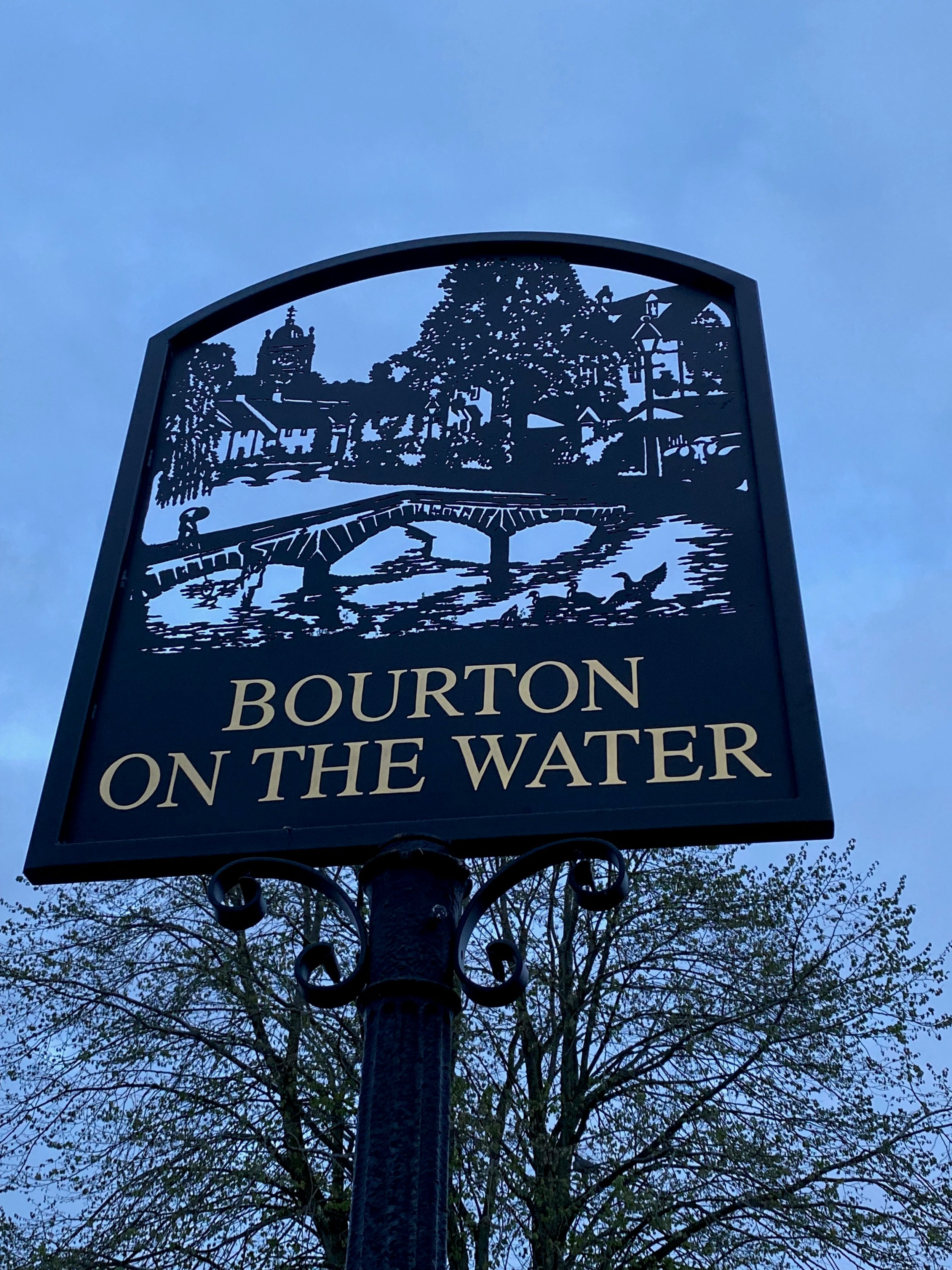A decorative sign for Burlington on the Water with a detailed cutout scene of a park, water, and buildings, mounted on a black metal post against a blue sky with trees in the background.