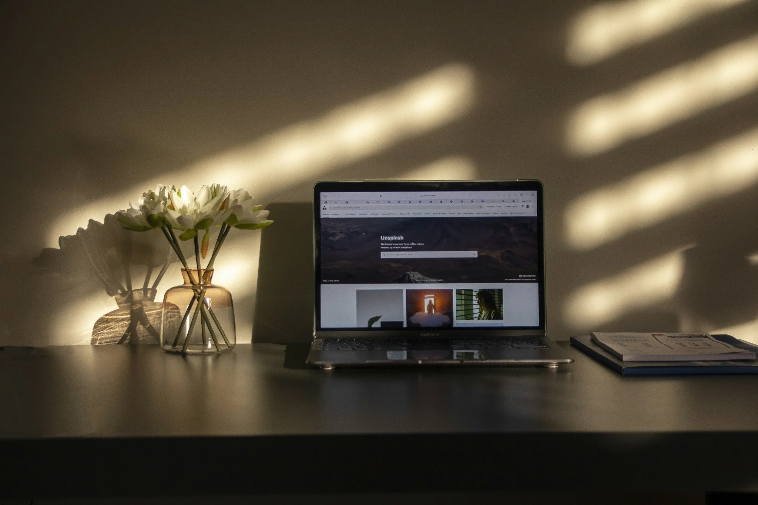 A laptop on a desk with a flowers vase on the left, and a closed notebook or journal on the right. The background shows a wall with light and shadow patterns.