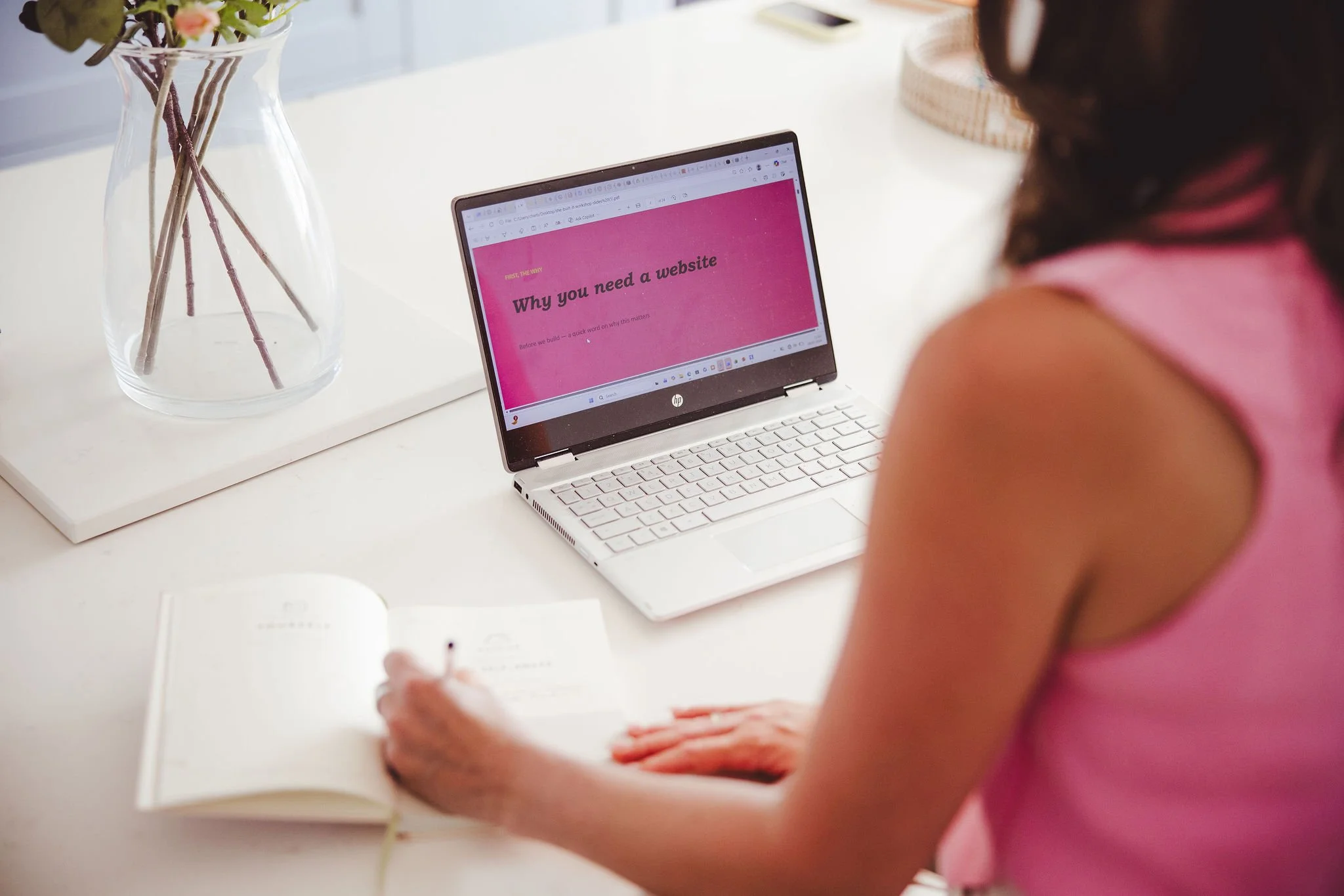 A person wearing a pink top sitting at a white desk, working on a laptop with a pink slide that says "Why you need a website" on the screen. A large glass vase with flowers is positioned to the left on the desk, and a notebook is open in front of the person.