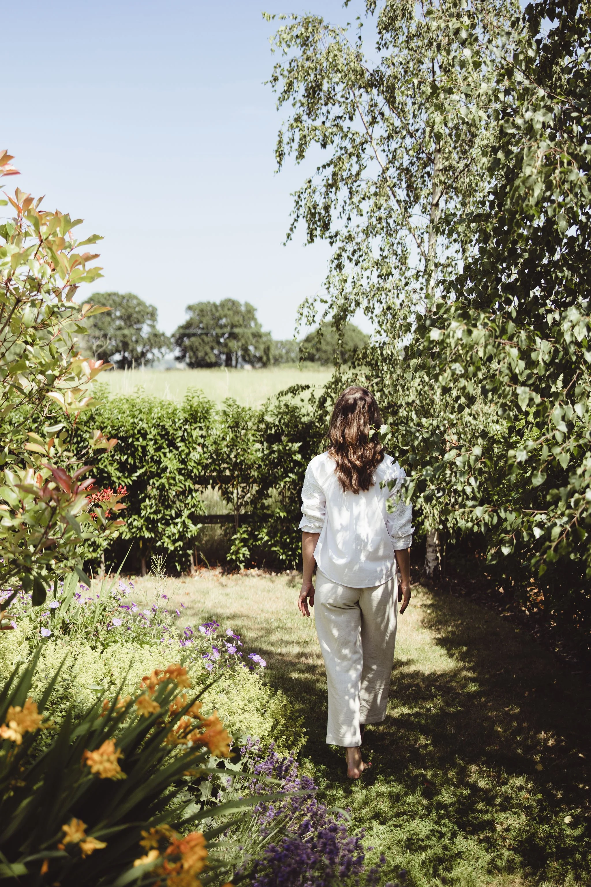 A woman with long brown hair, dressed in an off-white shirt and light-colored pants, walks barefoot along a garden path surrounded by various green plants and flowers, with trees and a bright blue sky in the background.