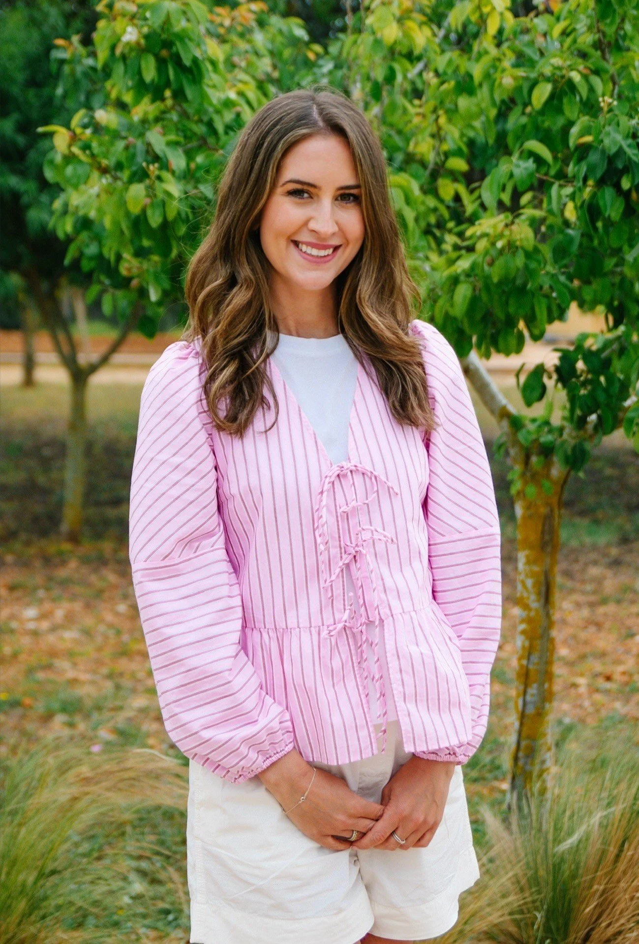 A woman with wavy brown hair, smiling, wearing a pink and white striped blouse and white shorts, standing outdoors with green trees and grass in the background.