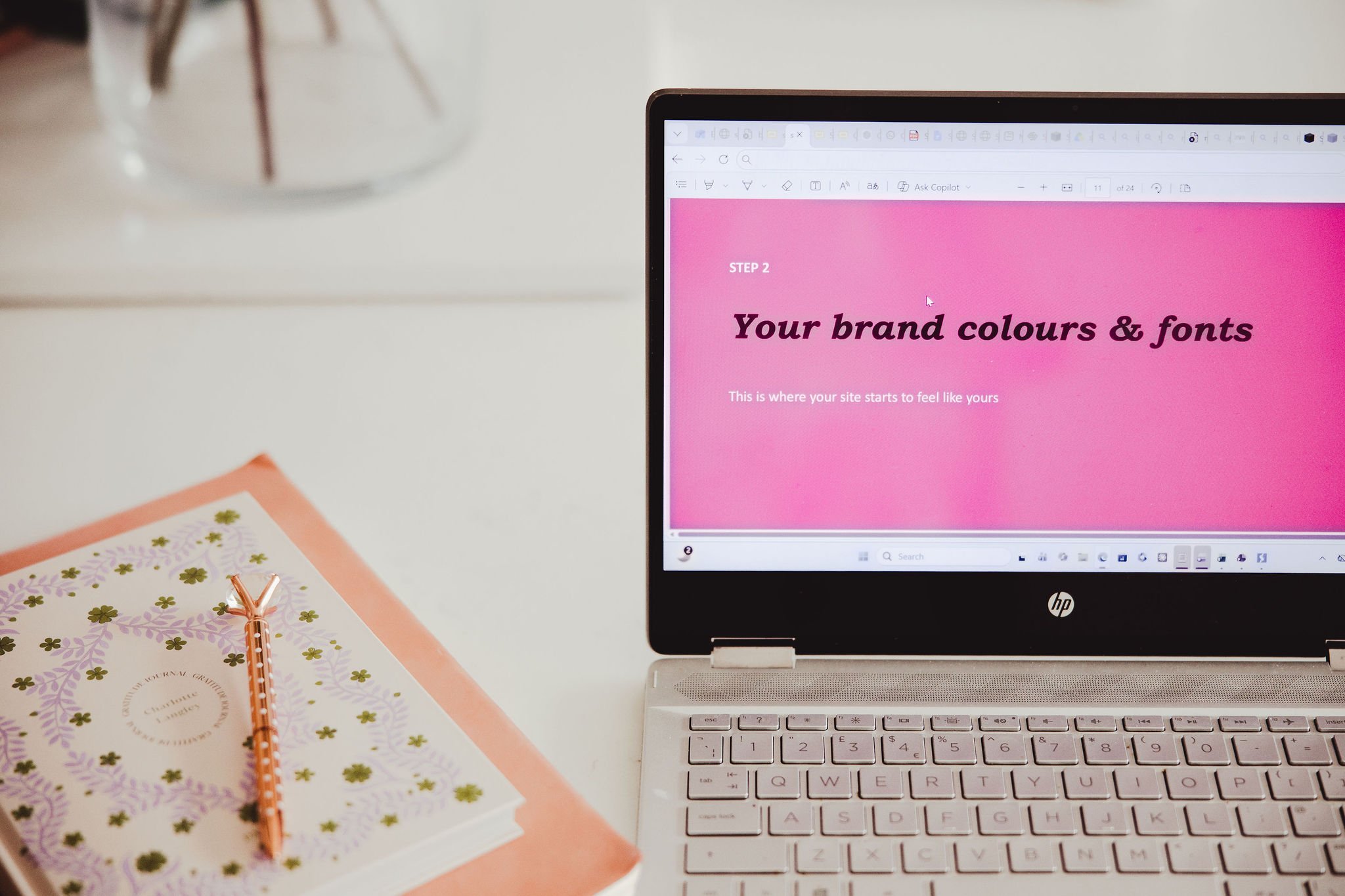 Open HP laptop displaying a webpage with pink background and black text, a decorated notebook with a rose gold pen on top is on a white desk, and a clear glass vase with a plant is in the background.