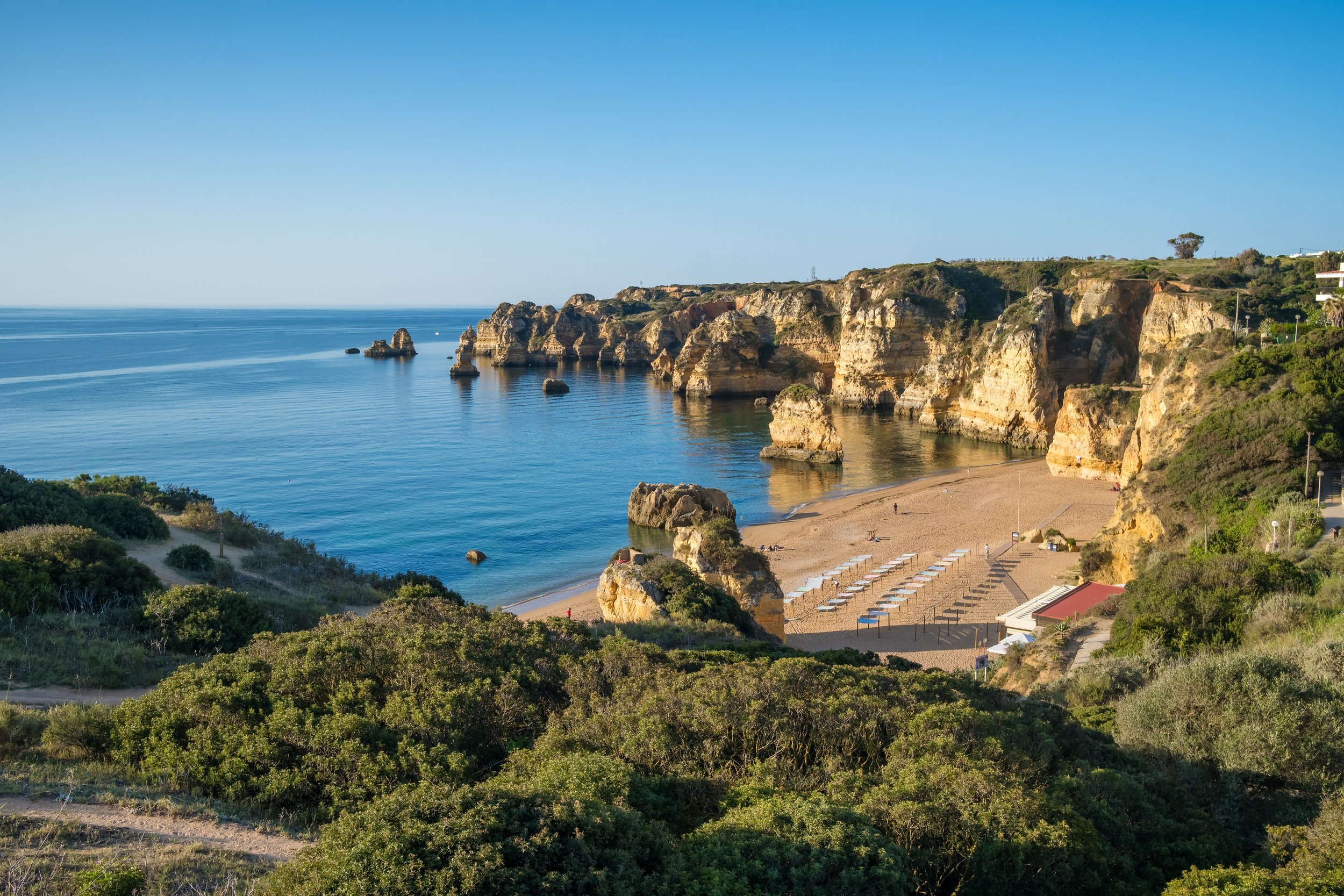 Cliffs overlooking a beach with sun umbrellas, a sandy shoreline, and calm ocean waters under a clear blue sky.
