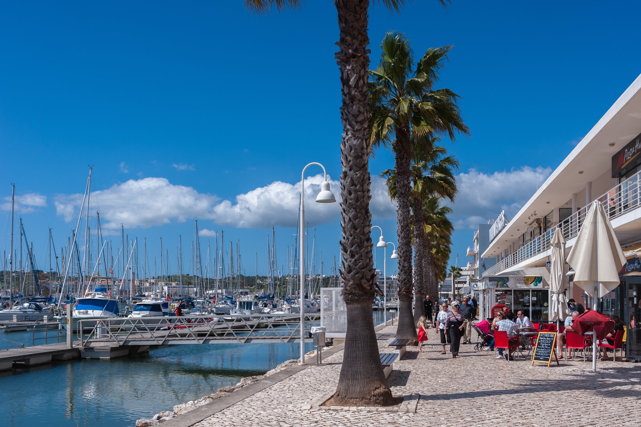 Marina with boats docked at the pier, palm trees lining the promenade, and a building with outdoor seating and shops against a bright blue sky with few clouds.