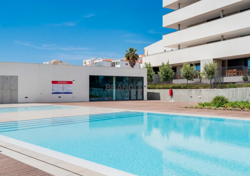 Outdoor swimming pool area with modern buildings in the background, clear blue sky, and some greenery.