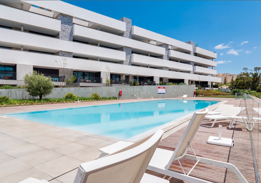 Outdoor swimming pool with lounge chairs in front of a modern apartment complex on a sunny day.