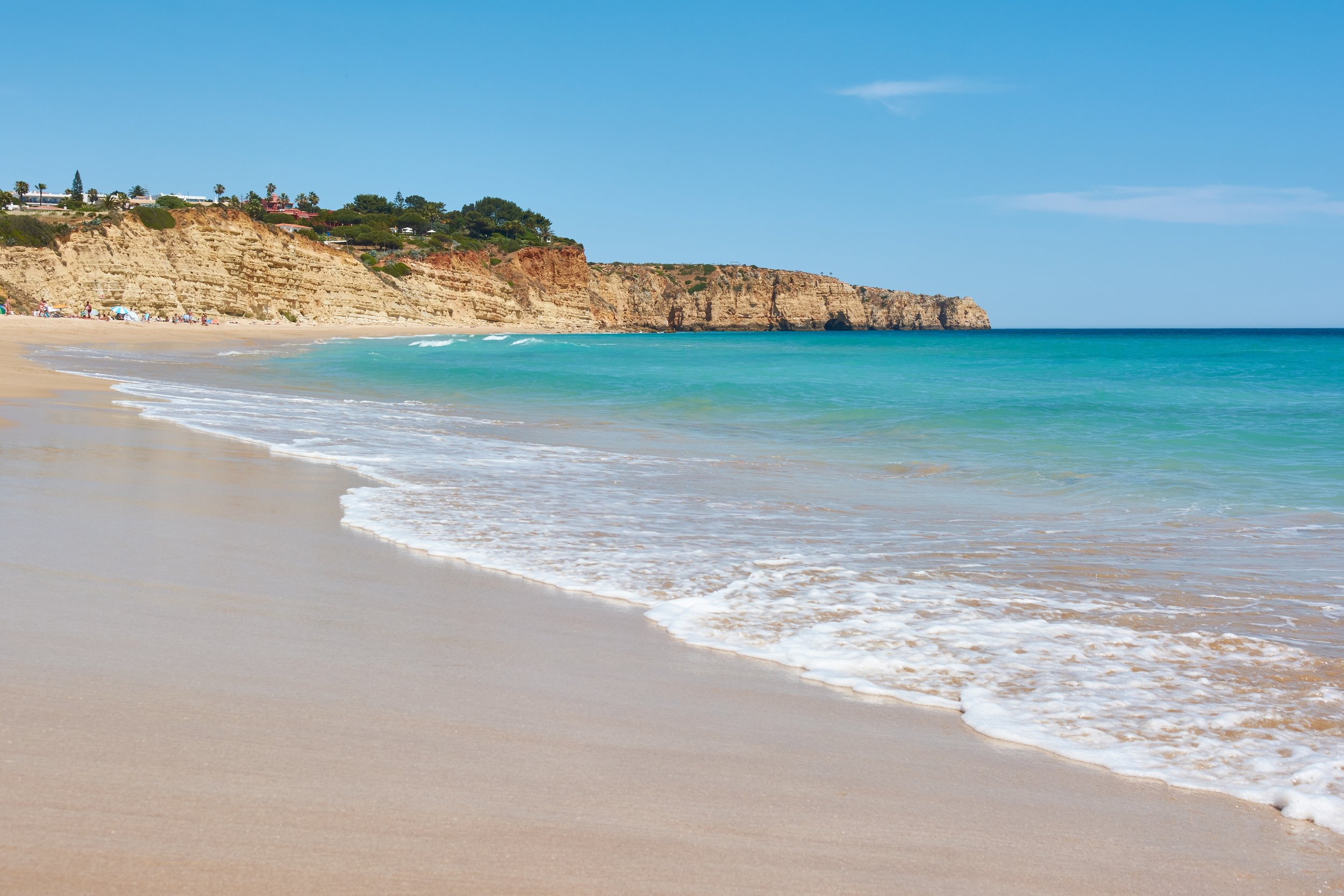 Beach with sandy shore, ocean waves, cliffs with vegetation and houses in the background, clear blue sky.