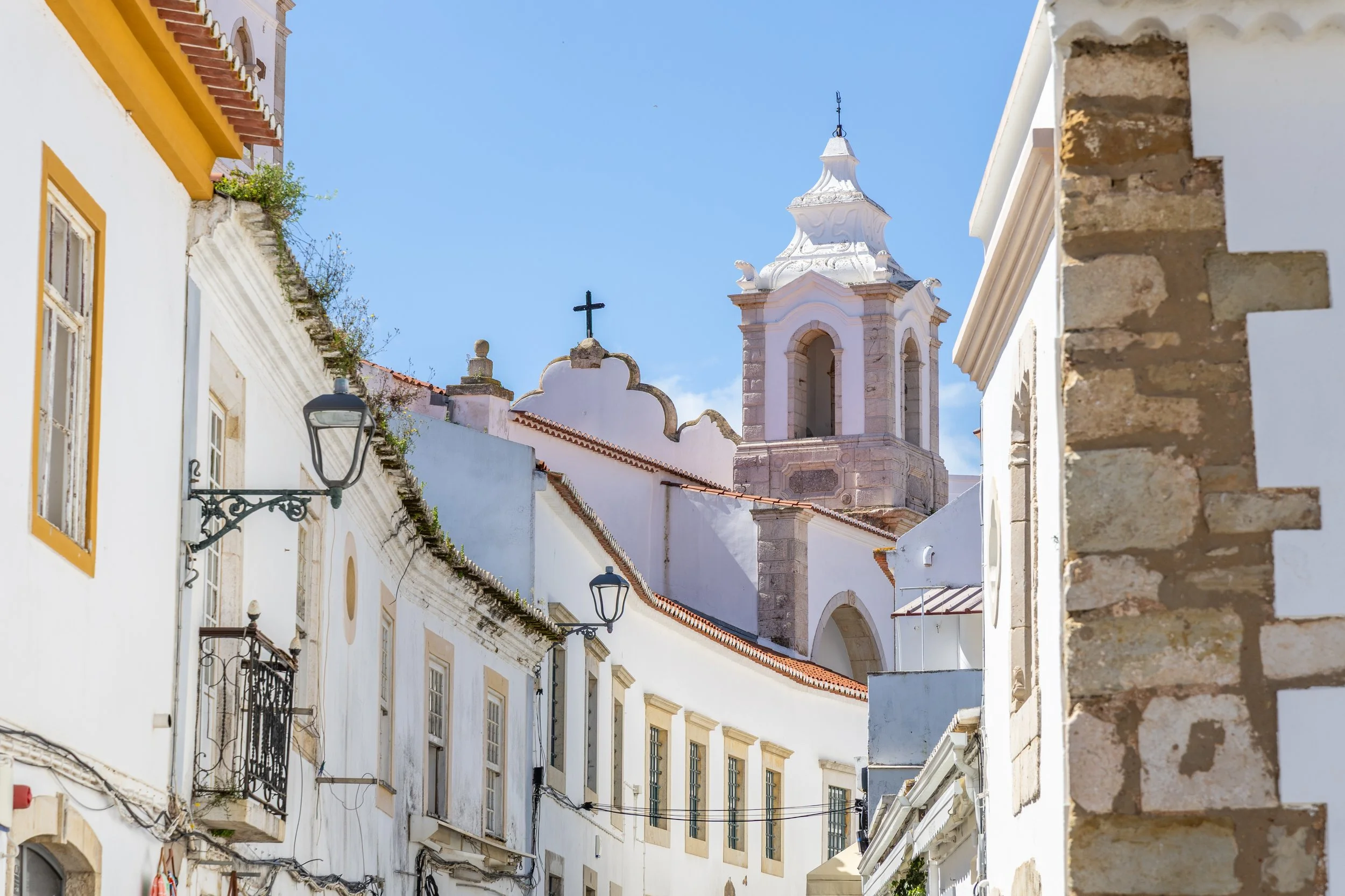 A narrow street with white buildings, black wrought-iron balconies, and a church with a tall bell tower and cross against a bright blue sky.