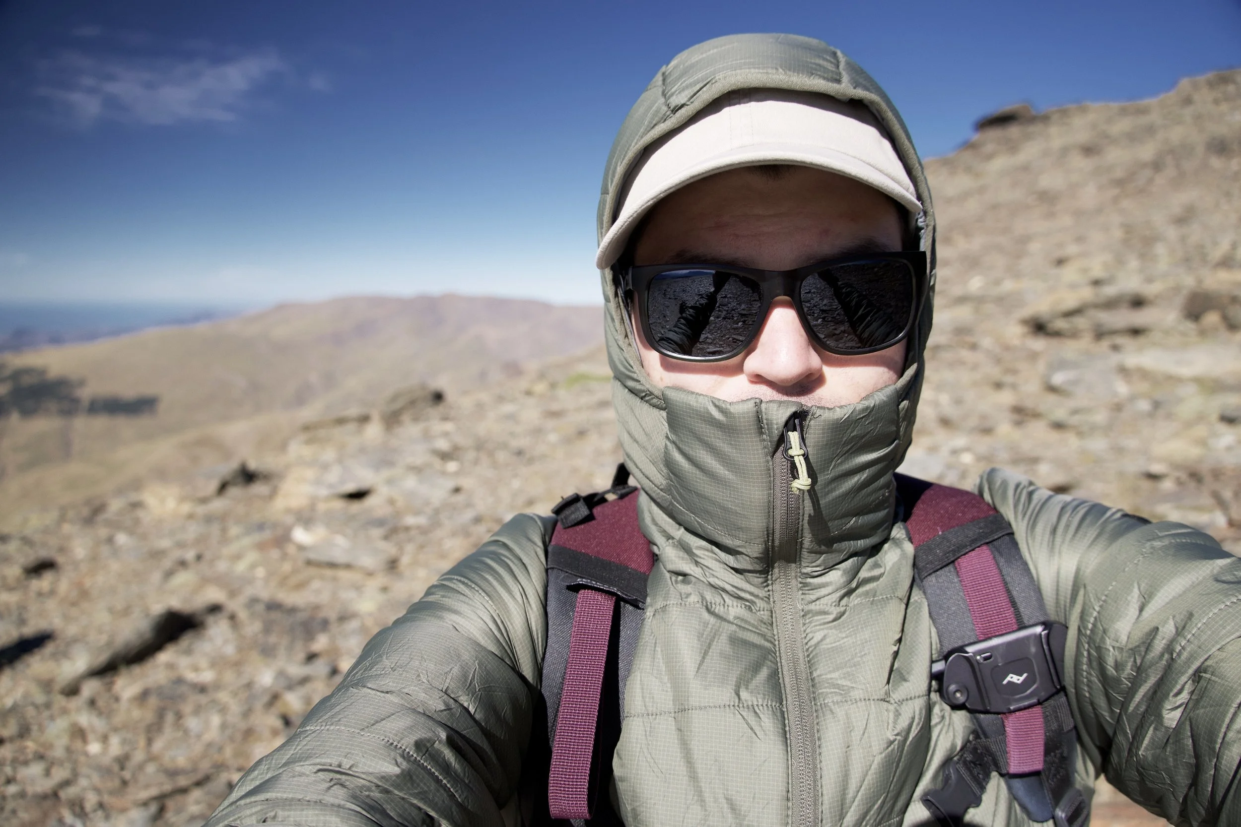 A person taking a selfie outdoors in a rocky, mountainous area wearing a light gray jacket, a beige cap, black sunglasses, and a purple backpack.