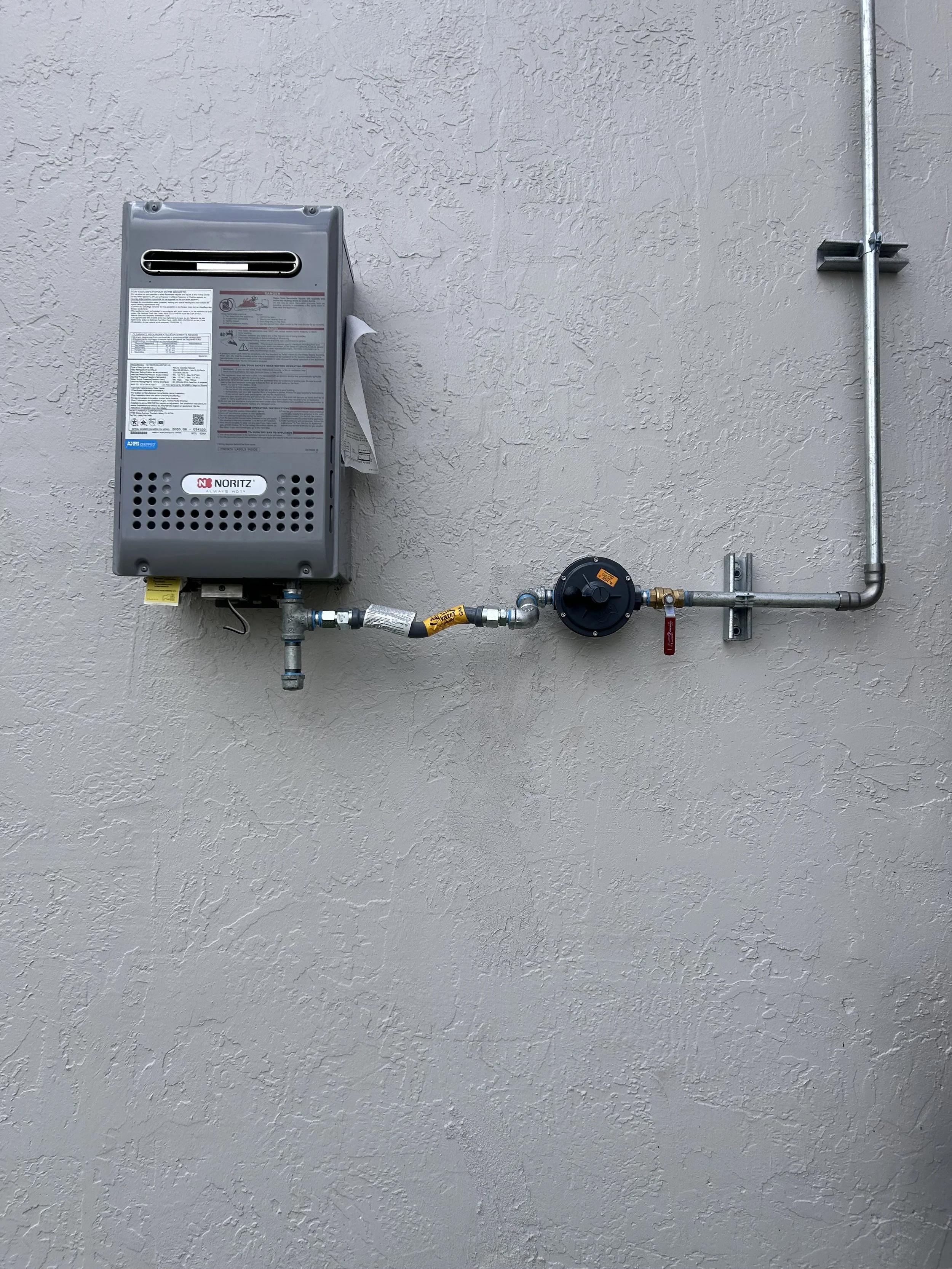A wall-mounted gas water heater with pipes, valves, and a control panel, installed on a stucco-textured beige wall.