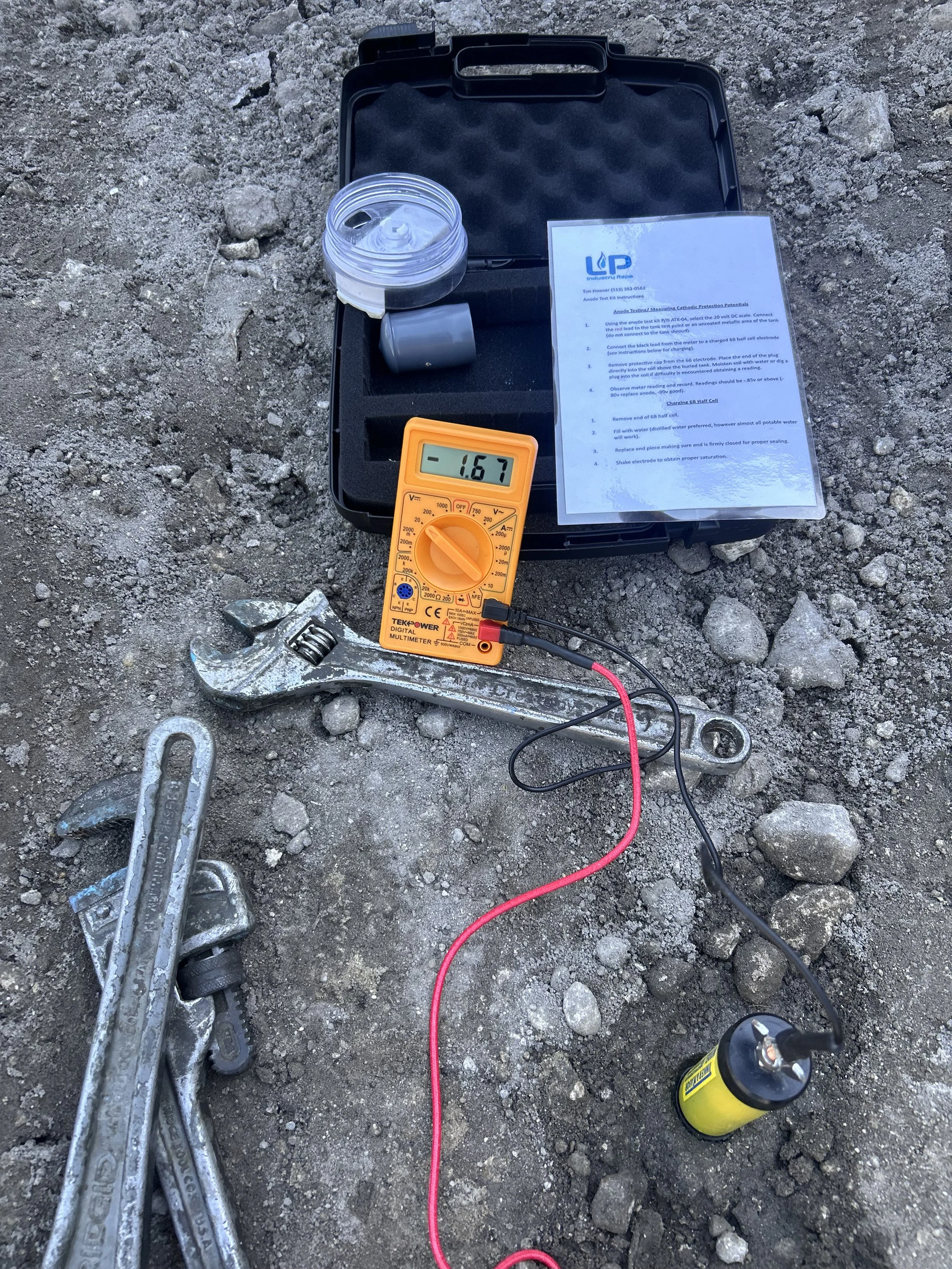 Tools and equipment for electrical testing on rocky ground, including a multimeter, a protective cap, a test kit, a wrench, and a flashlight, with instructions sheet.