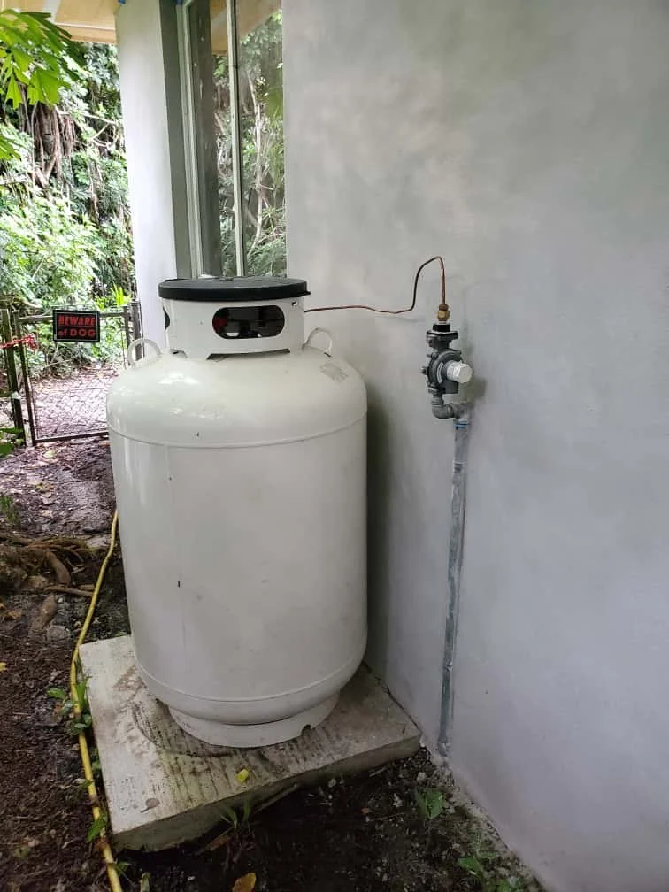 A white propane tank connected to a pipe and regulator outside a house near a garden with trees and a 'Beware of Dog' sign.