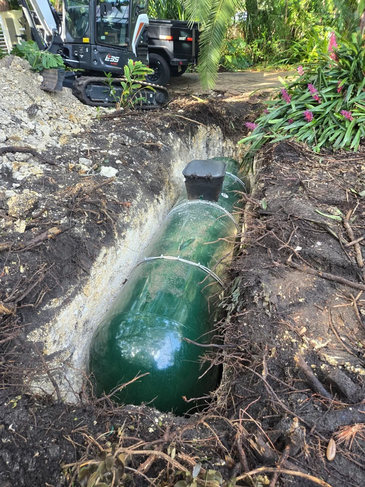A green underground Propane tank is being installed in a trench, with soil and roots surrounding it. A small construction excavator is nearby in a lush, garden-like area.