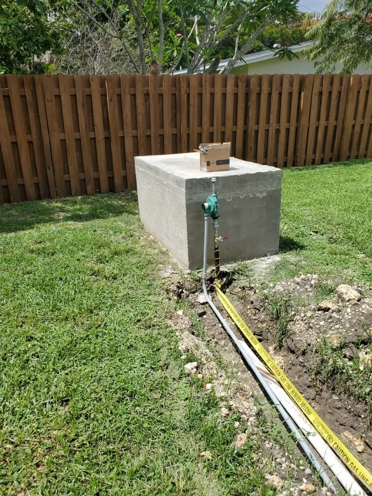 A concrete pedestal with a Generator, a Second Stage Regulator, and a pipe extending from it, set in a grassy backyard with a wood fence and trees in the background.