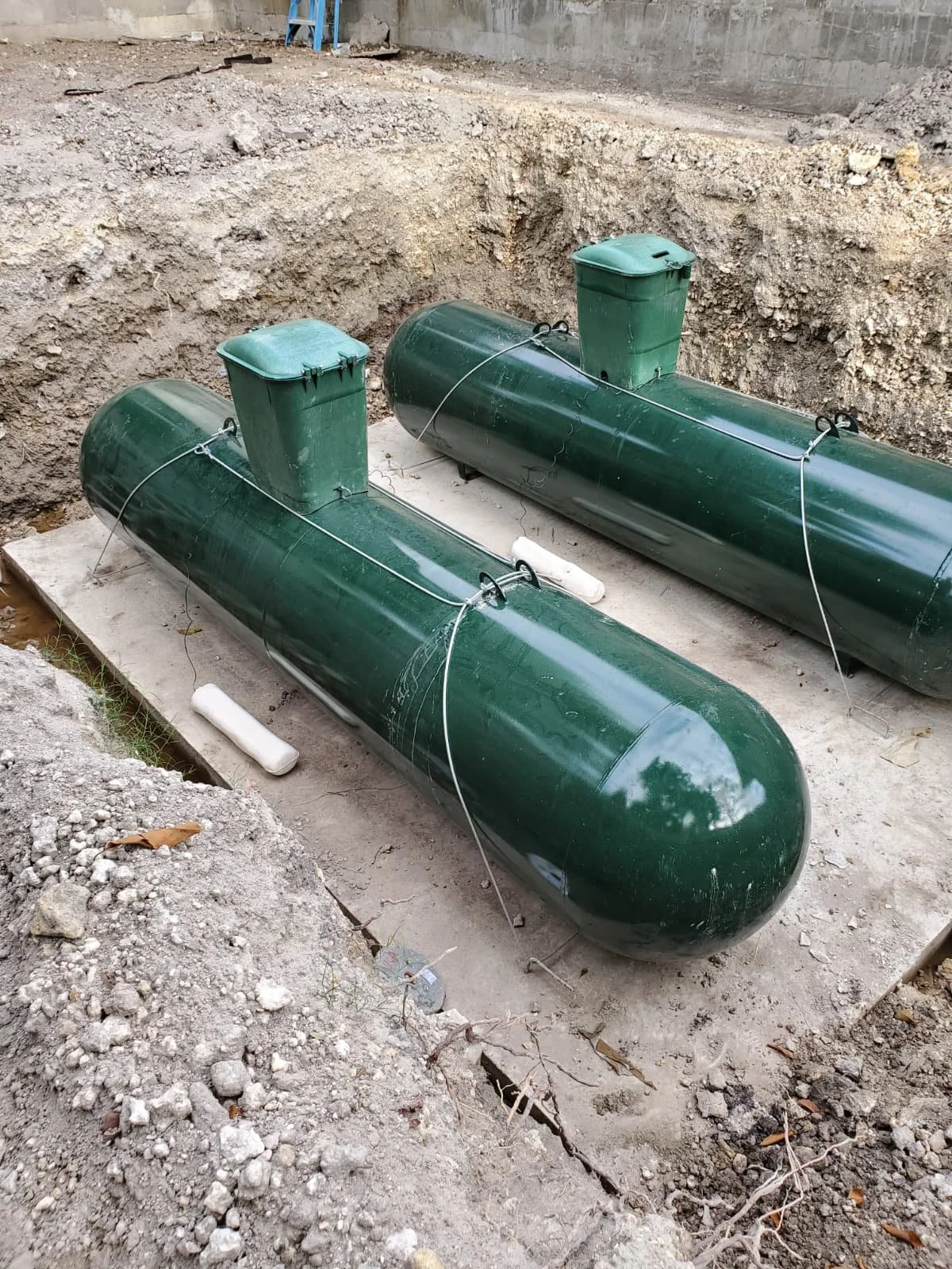 Two large green underground storage tanks with safety valves, installed in a construction site with dirt and concrete surroundings.