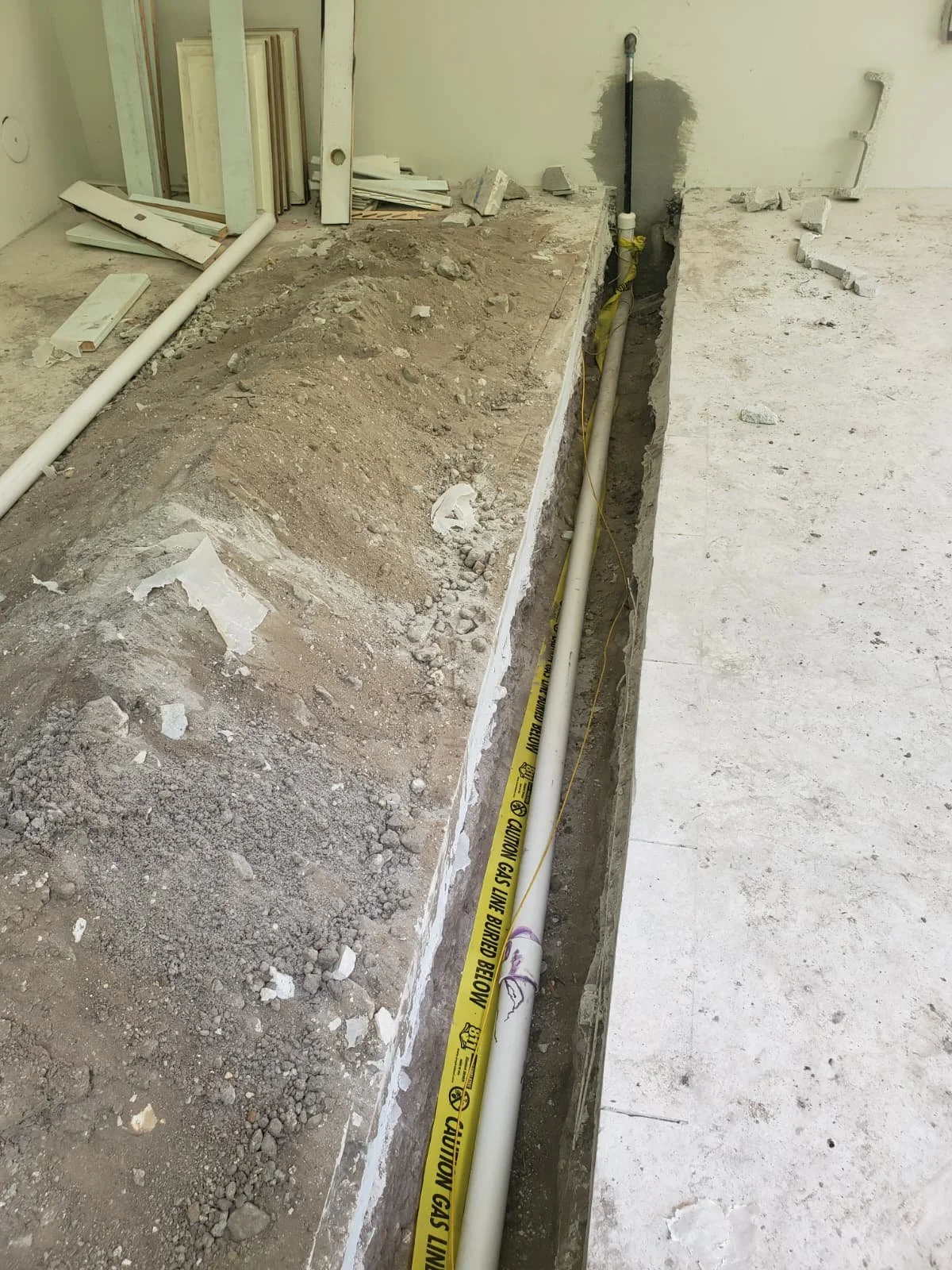 Construction site with exposed plumbing and gas pipes running along a deep trench in the floor, and some construction materials piled to the side.