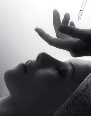 Close-up of a person lying down with their face and profile visible, a hand gently holding a dropper above their face, in a black and white photograph.