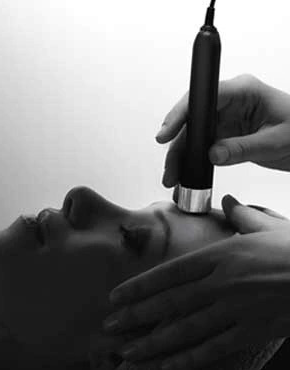 A person receiving a facial treatment with a small device held by a technician, close-up view in black and white.