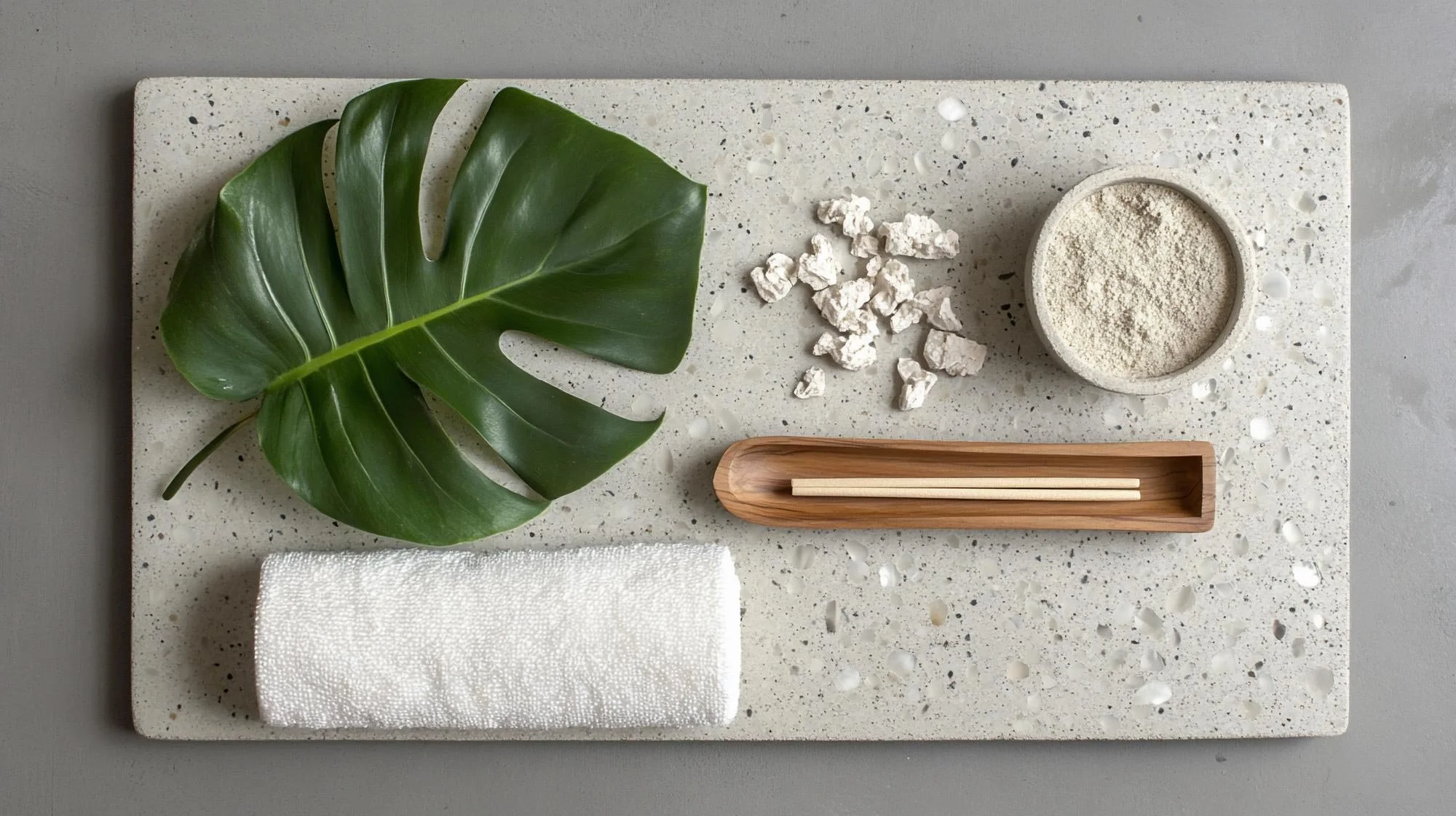 A flat lay of a spa or wellness setting featuring large green monstera leaves, a white textured towel, a wooden tray with chopsticks, a small bowl of white powder, and some white rocks on a speckled white stone surface, next to a gray wall.