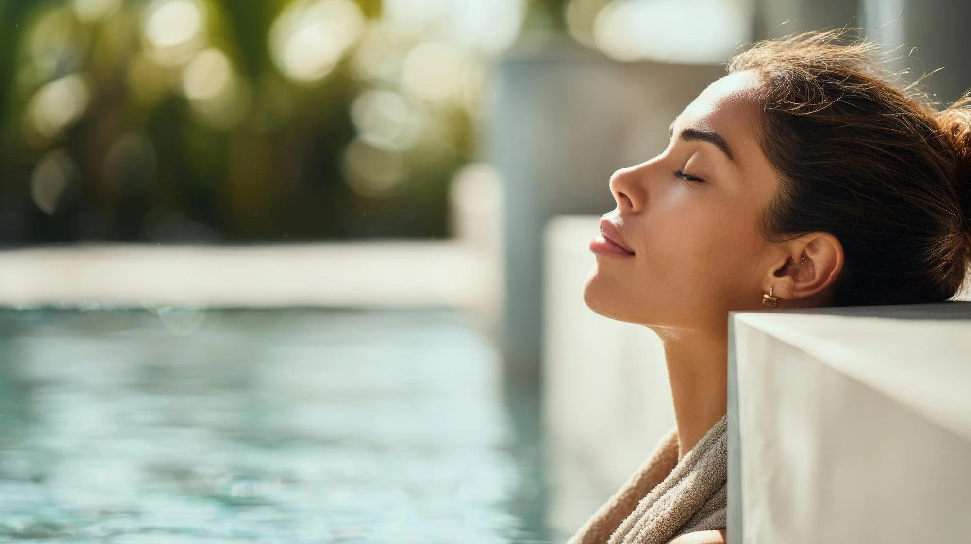 A young woman with closed eyes and relaxed expression rests her head on the edge of a swimming pool, with sunlight shining on her face and water in the background.