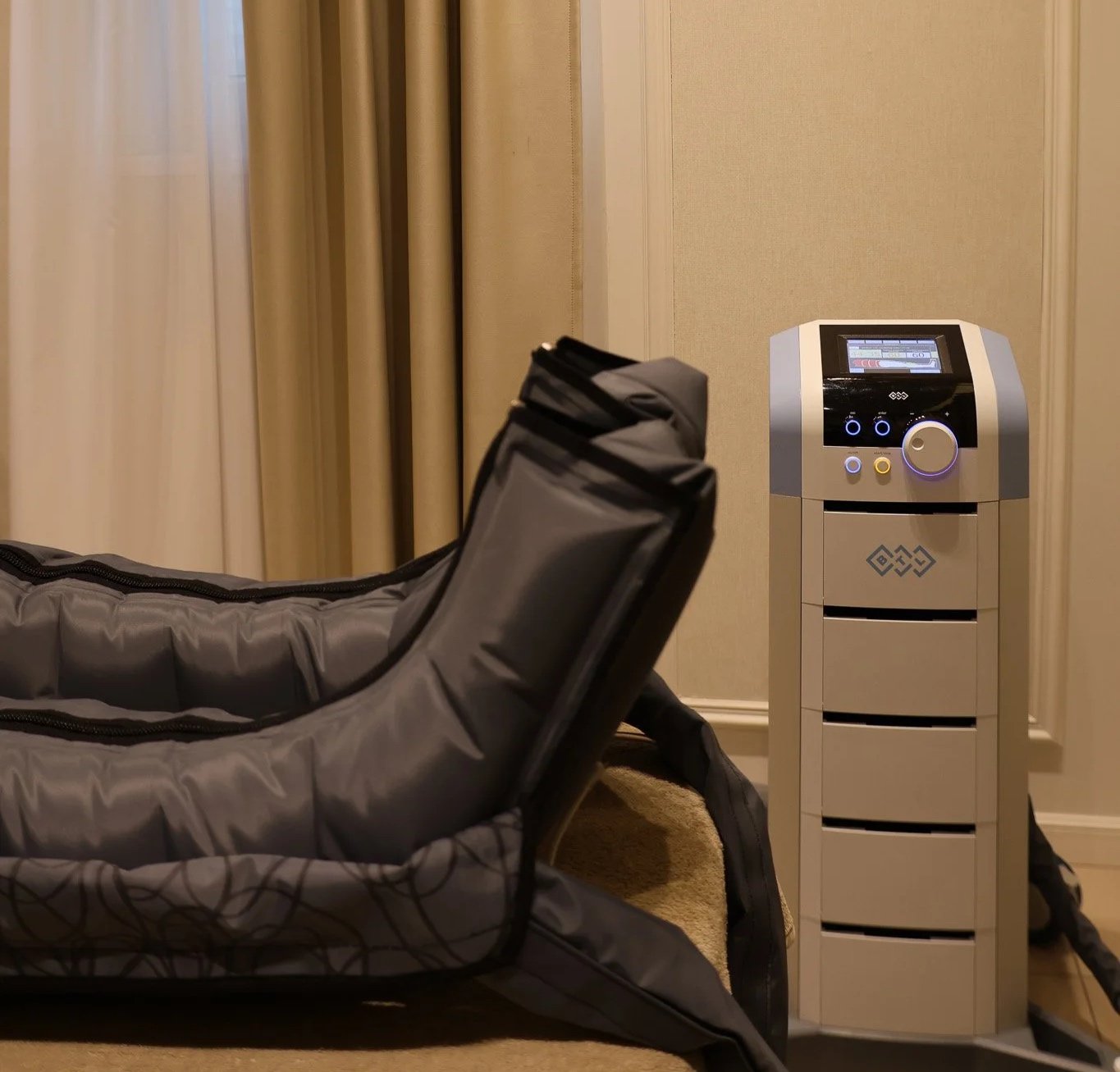 A massage or spa room with a padded massage table and a nearby machine, possibly for hot stones or massage tools, set against beige curtains and wall.