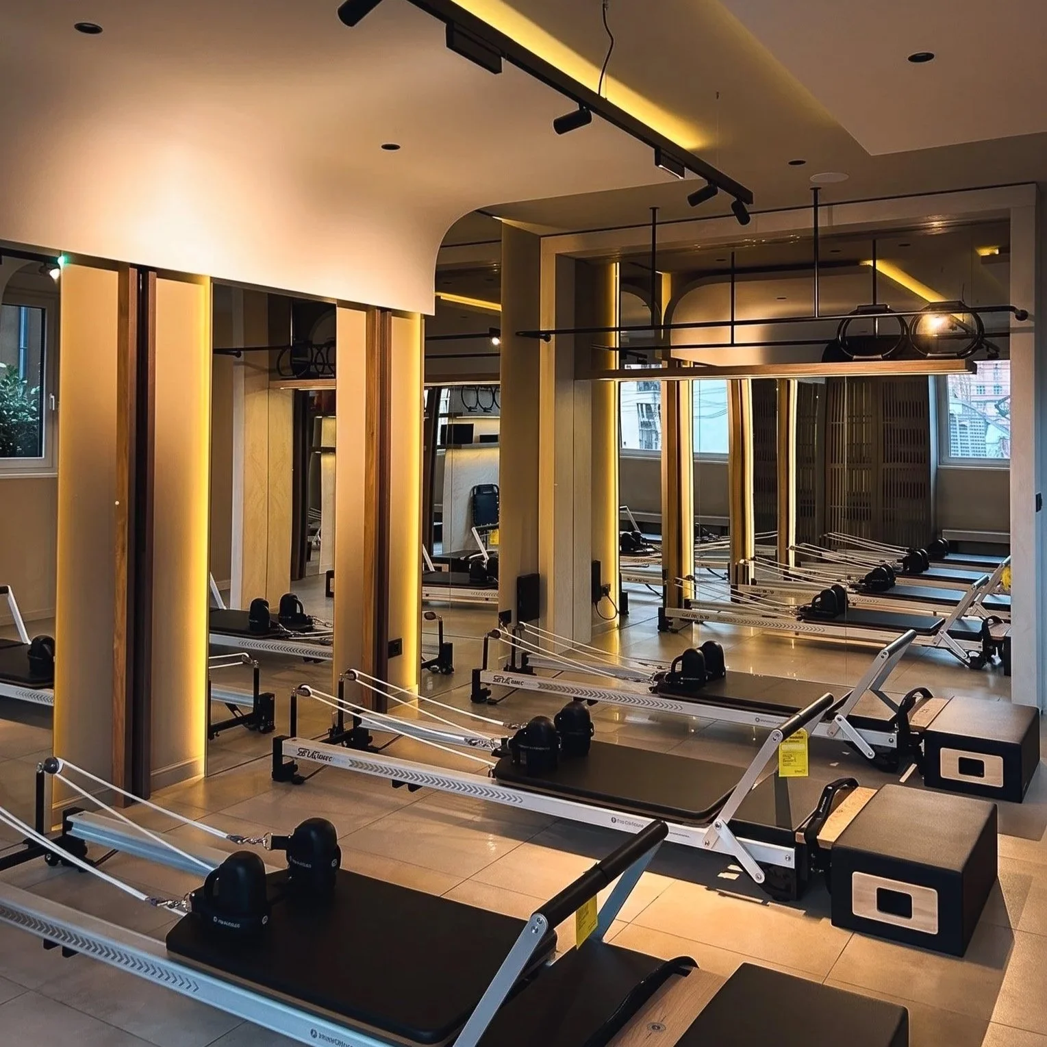 Fitness studio with reformer Pilates machines arranged in rows, illuminated by warm lighting, with mirrors on the walls and windows showing city buildings outside.