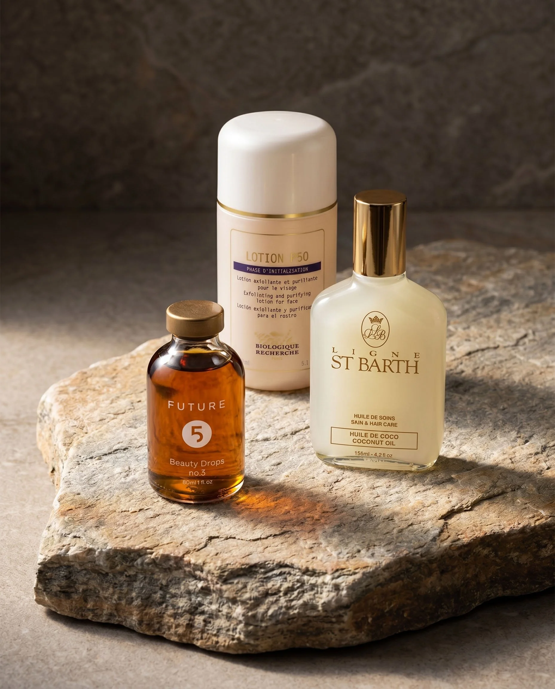 Three skincare or cosmetic bottles placed on a rough stone surface with a dark gray background. The bottles vary in size and color, with one amber-colored bottle, a tall white bottle with gold accents, and a clear bottle with a gold cap.