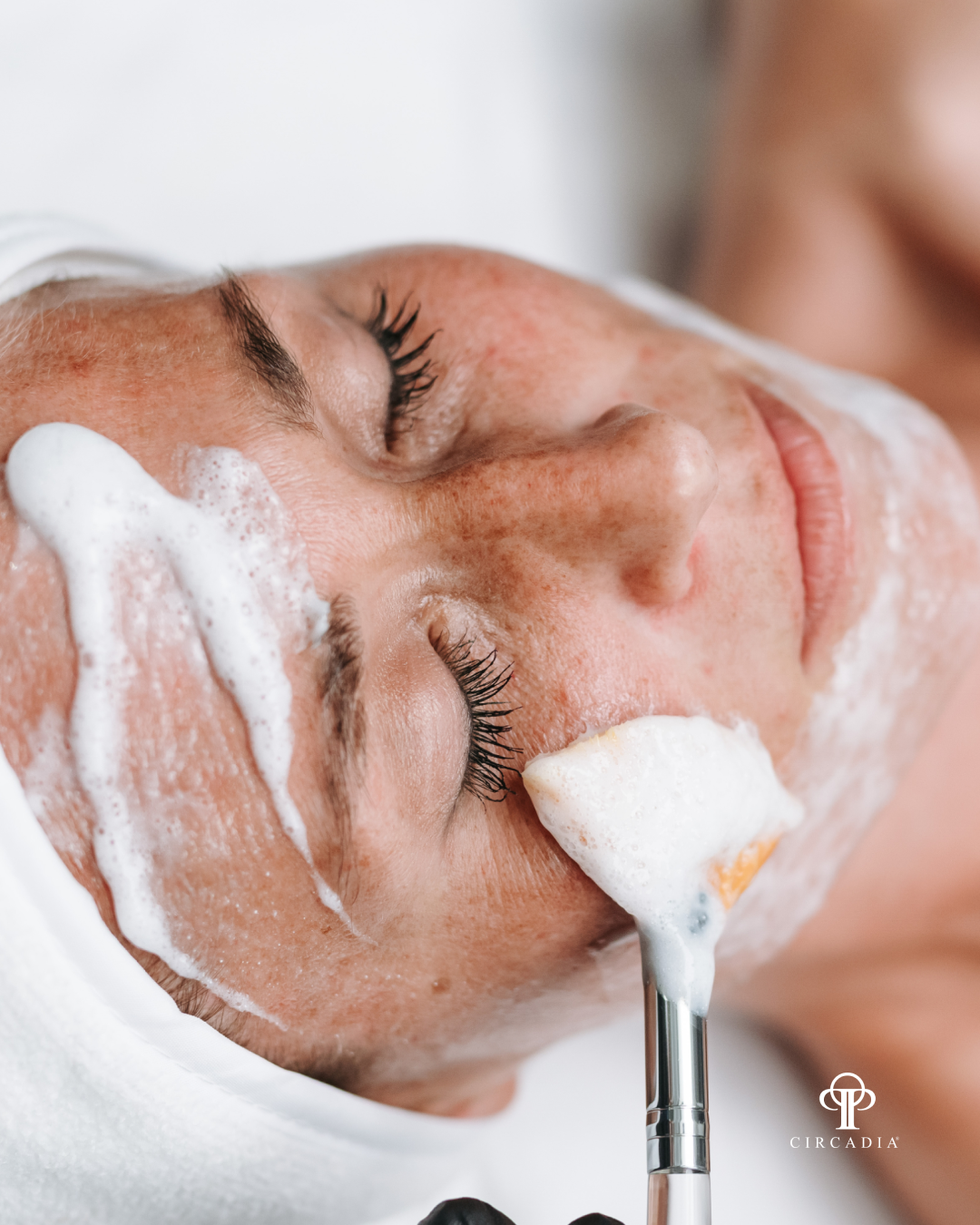 A woman receiving a facial treatment, with foam and a brush on her face, lying with eyes closed.