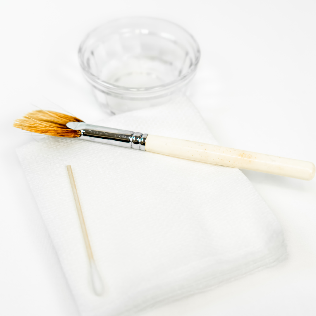 Facial brush, clear container with water, white applicator stick, and a paper towel on a white surface.