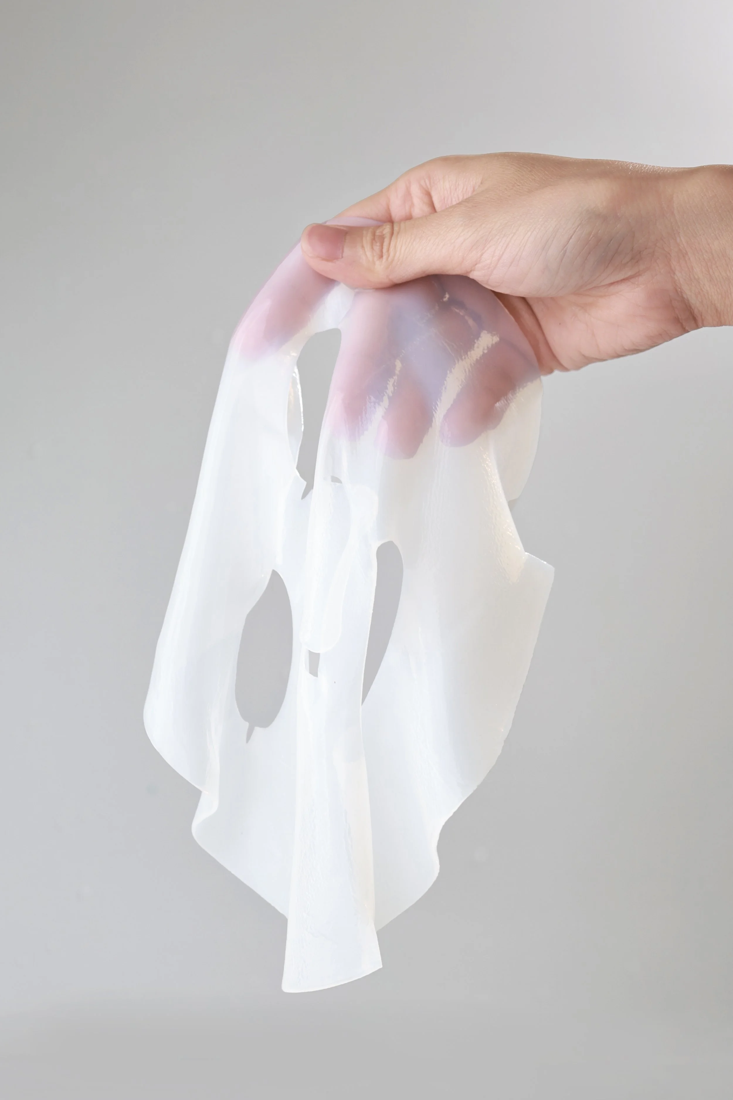 A person holding a semi-translucent, white, facial mask with a light gray background.