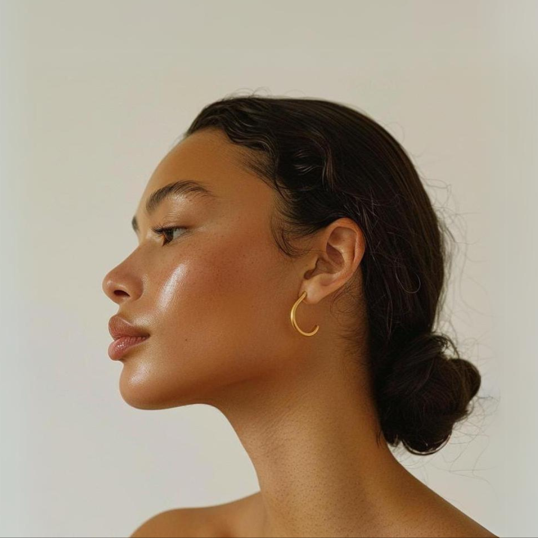 Side profile of a woman with sleek dark hair in a low bun, wearing a gold hoop earring, with glowing skin and a neutral background with glowing skin.