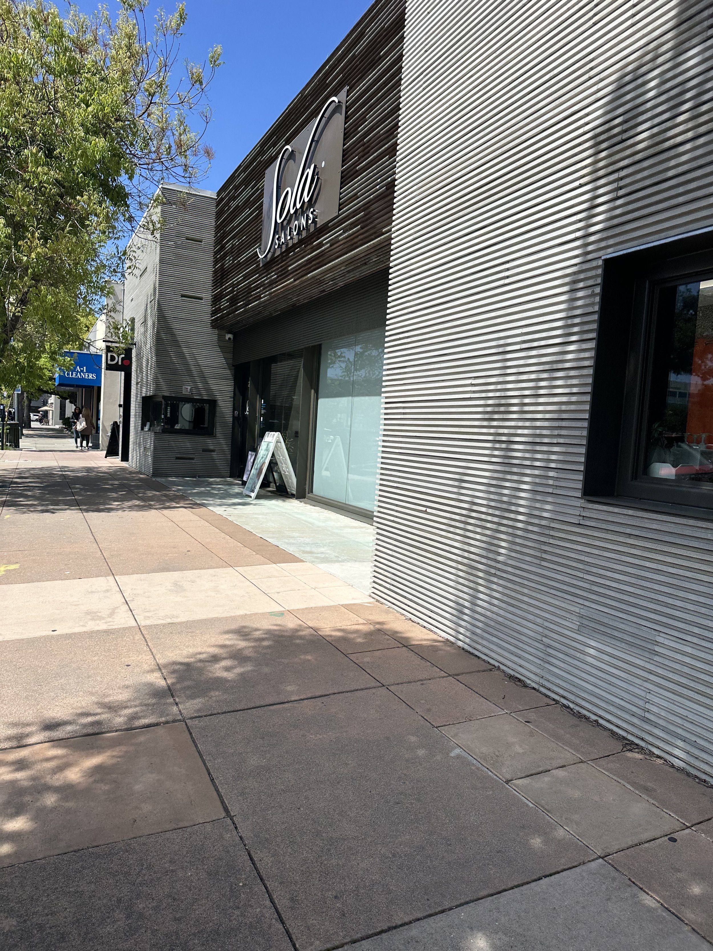 Sola Salon Burlingame, CA.  A street scene with a modern building featuring metal siding and large glass windows. There are trees and a sidewalk with pedestrians, as well as various storefront signs, including one for a dry cleaner and a salon.