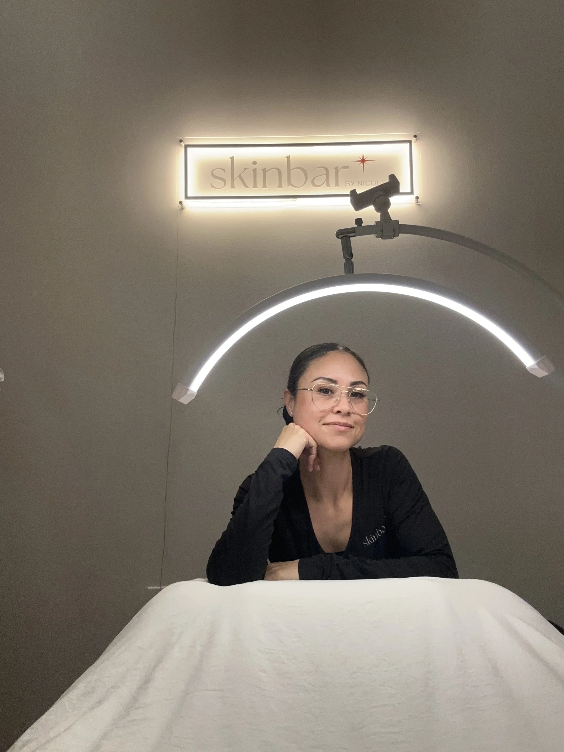 A woman sitting at a table in a spa or skincare clinic, with a neon sign above her that says 'skinbar by Nicole.' She has dark hair, glasses, and is wearing a black top.