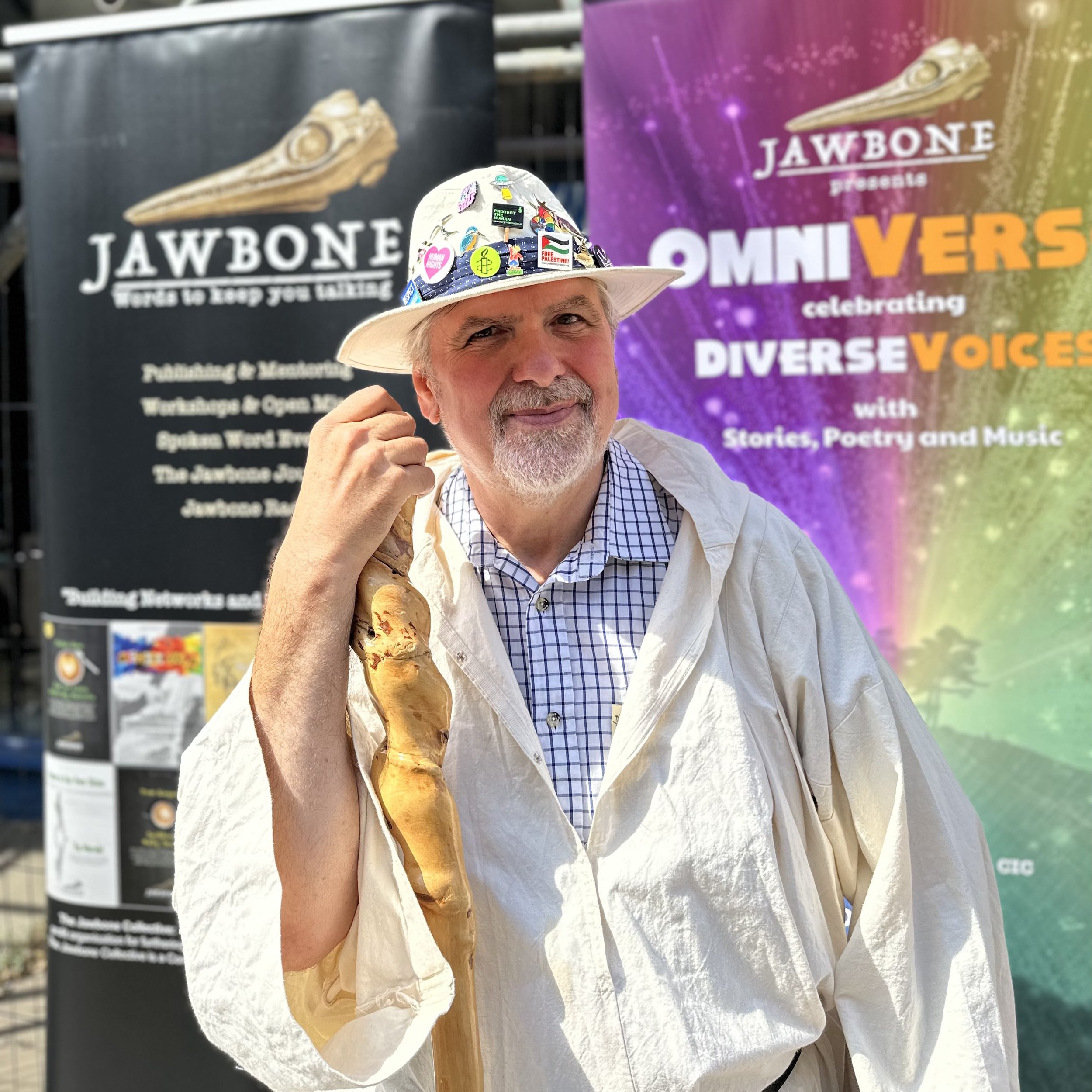 A man wearing a white hat decorated with various colorful pins and badges, holding an ice cream cone, standing in front of banners promoting JAWBONE and Omniverse celebrating diverse voices.