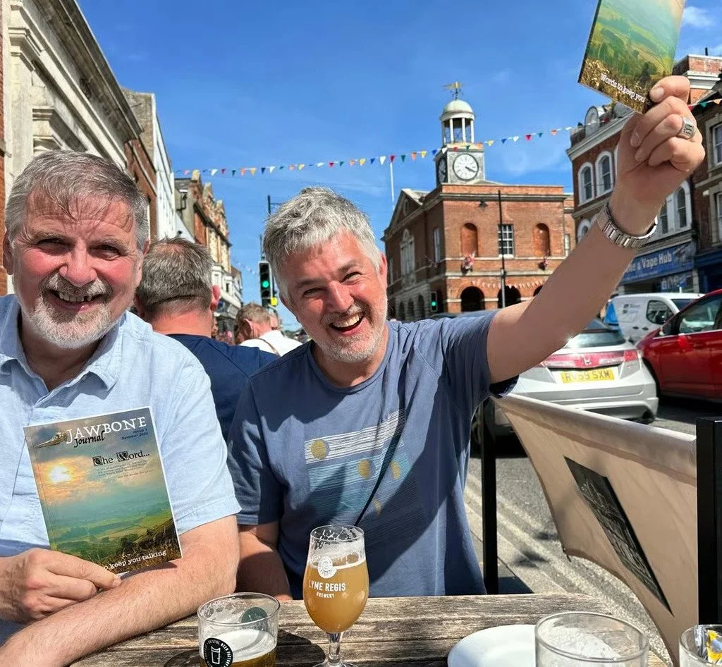 Two men sitting at an outdoor table on a city street, smiling and enjoying drinks, with a historic building with a clock tower and colorful bunting in the background.
