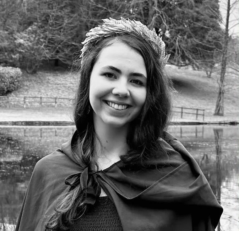 A young woman smiling with long wavy hair wearing a crown made of leaves and a cape, standing outdoors near a pond with trees in the background.
