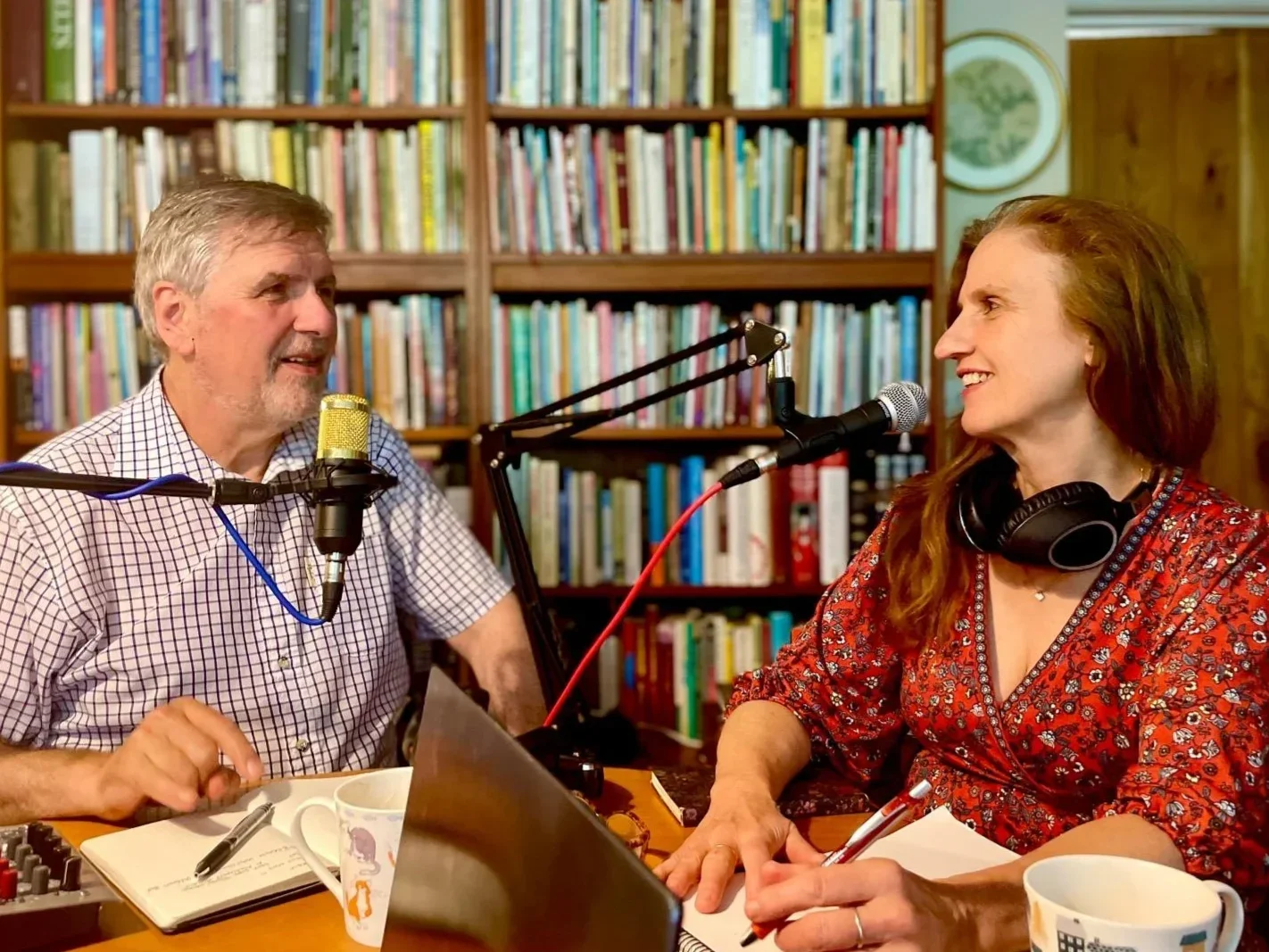 A man and woman sit at a table with microphones, recording a podcast or radio show in a room with a bookshelf filled with colorful books.