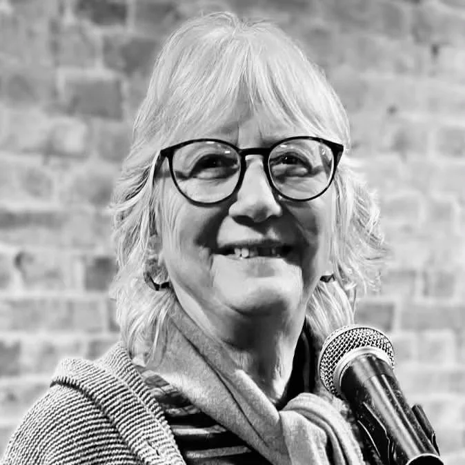 Black and white photo of a smiling woman with glasses and shoulder-length hair, standing in front of a brick wall with a microphone.