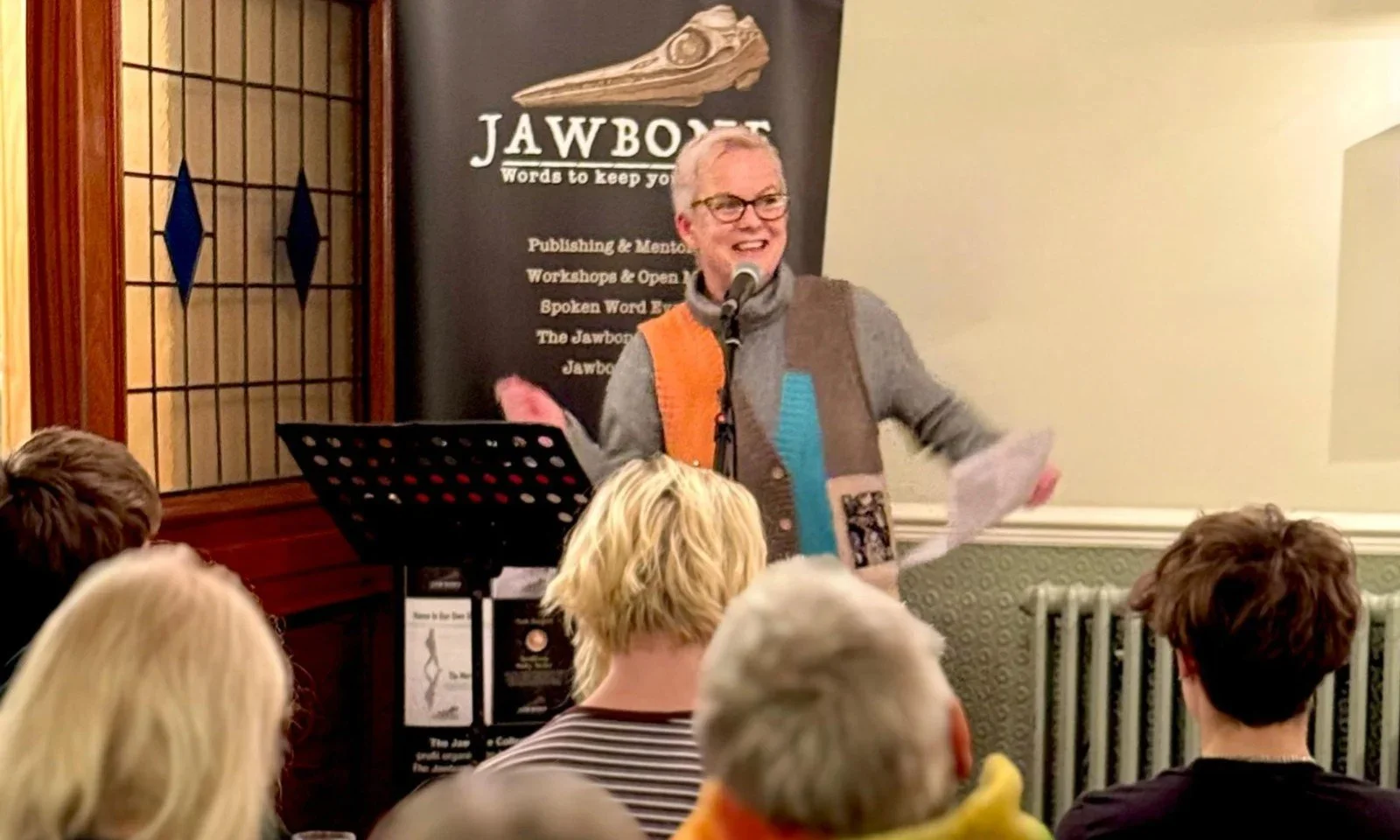 A person with glasses, short light-colored hair, wearing a colorful sweater vest over a gray shirt, is speaking at a microphone during a presentation or event. There are several audience members visible from behind, and a black banner with the word 'JAWBON' and an animal skull graphic is behind the speaker.