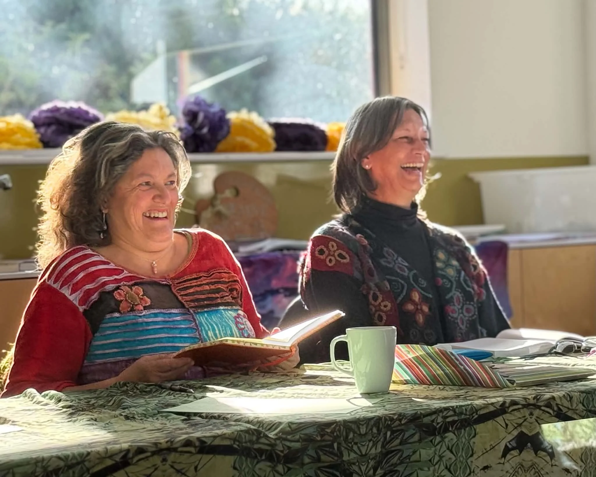 Two smiling women sitting at a table in a brightly lit room, with colorful blankets and a window with plants outside in the background.