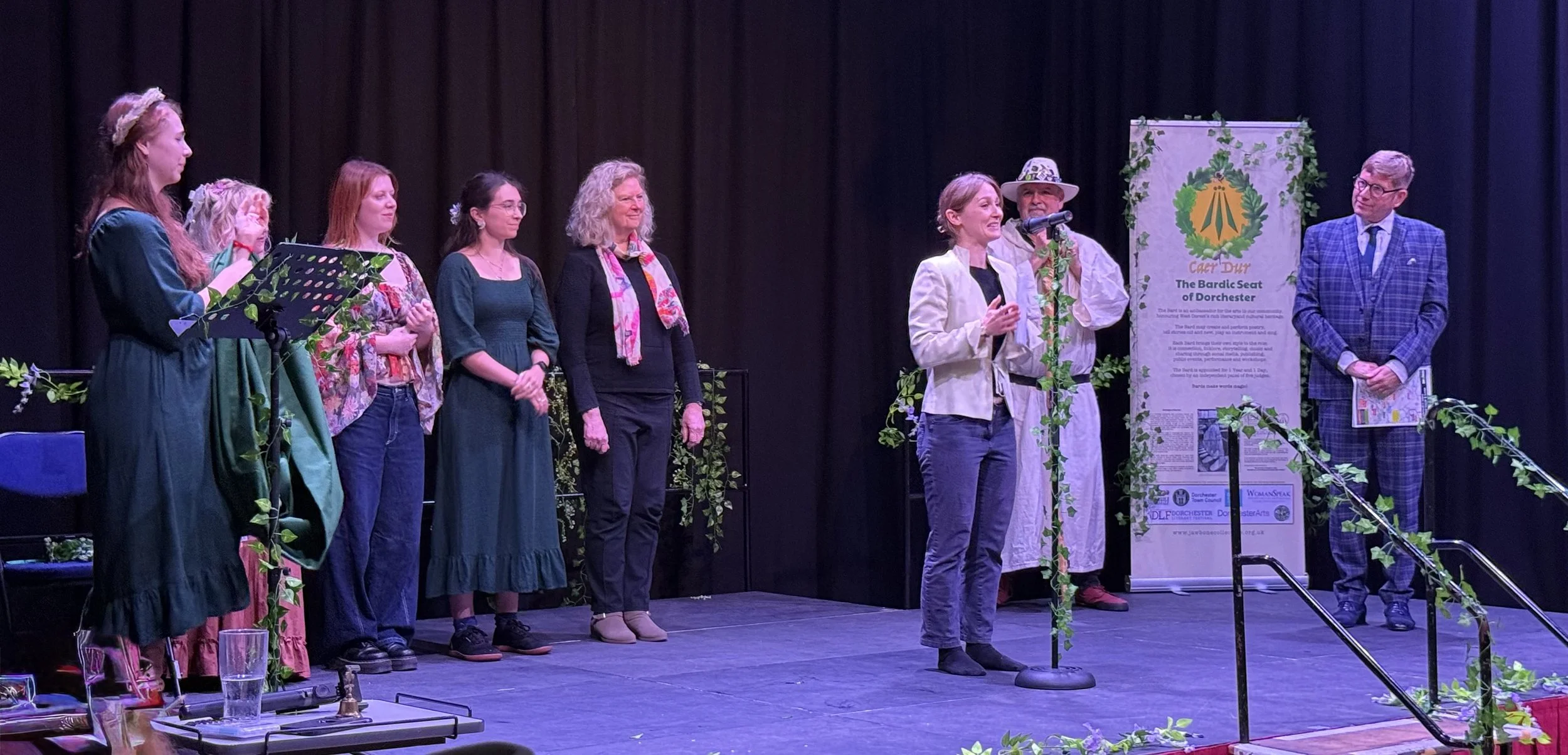 A group of eight women and one man standing on a stage decorated with green vines. One woman is speaking at a microphone, and a man in a plaid suit stands on the far right. There is a banner in the background with the words 'The Bardic Seat of Dorchester' and other text.