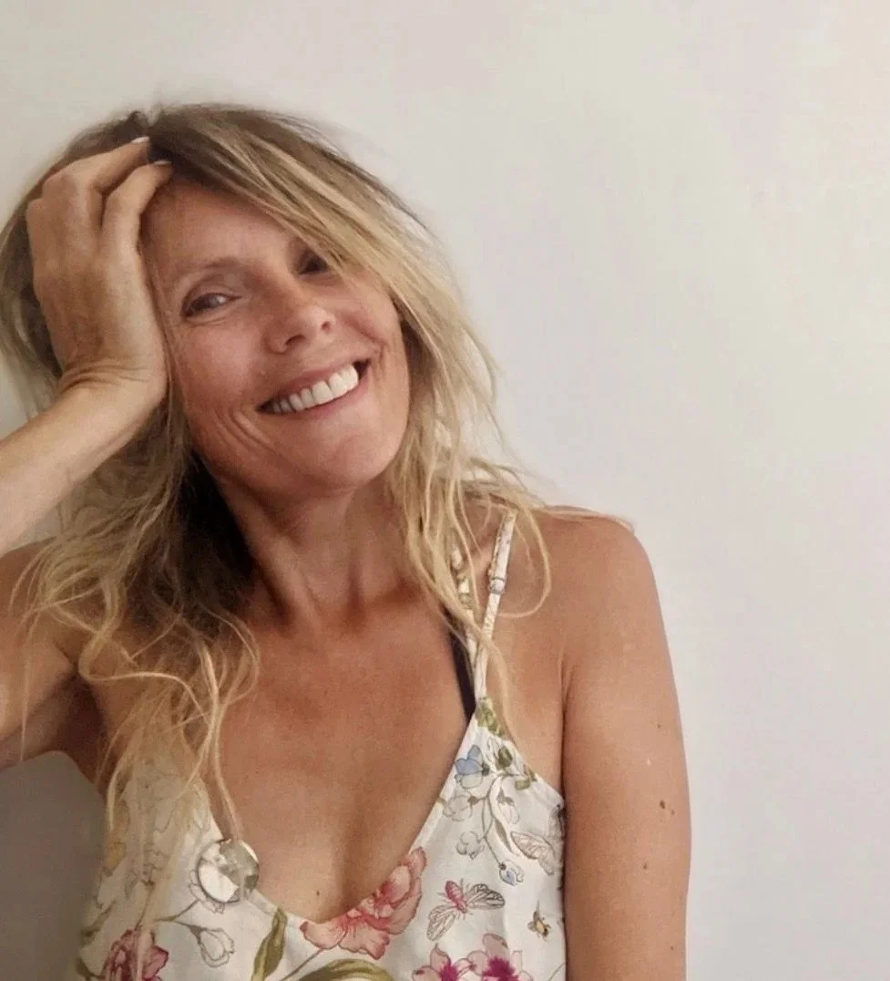 A woman with blonde wavy hair smiling and holding her hand on her head, wearing a floral spaghetti strap top, against a plain light-colored background.