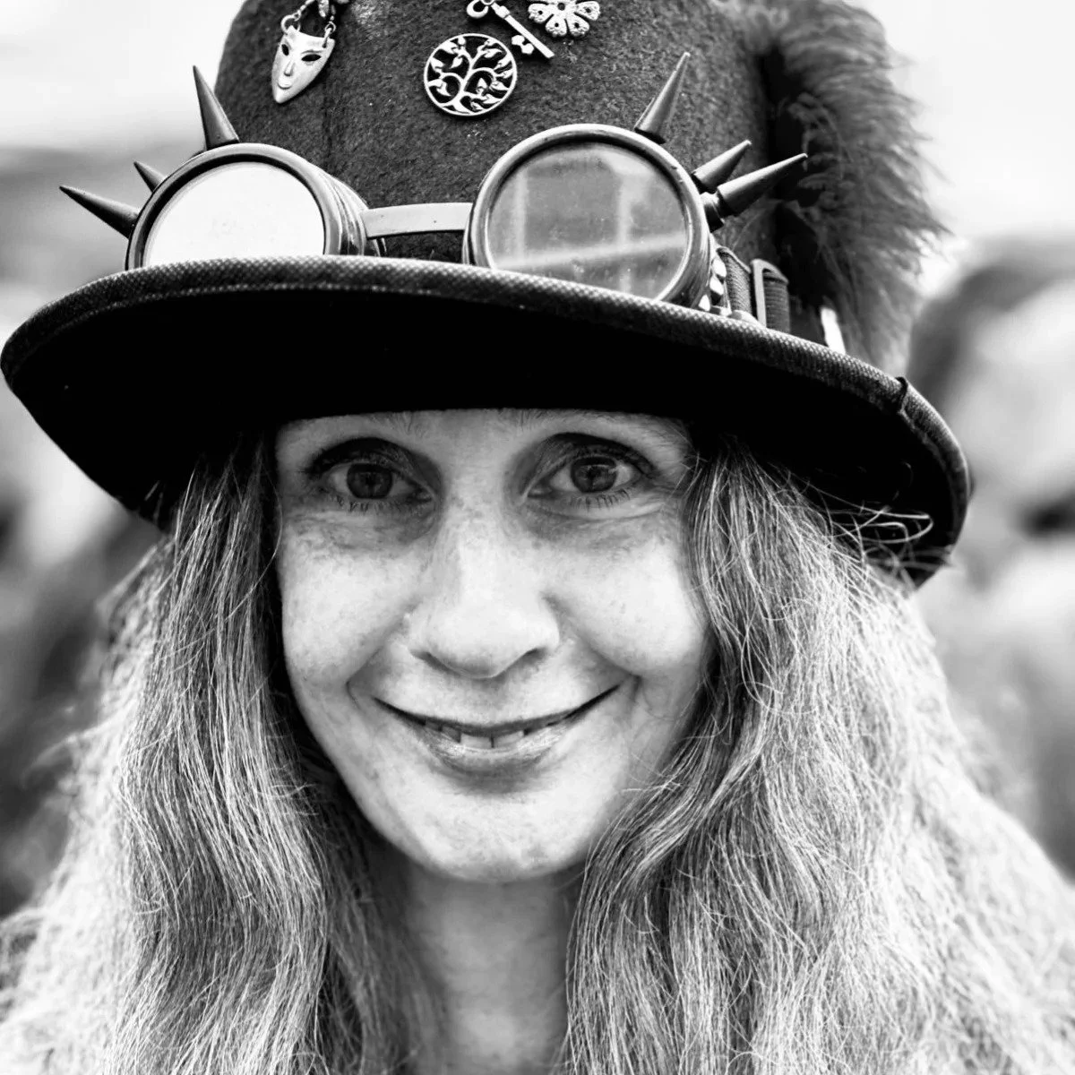 A woman with long hair wearing a hat with goggles, spikes, and pins, smiling at the camera in black and white.