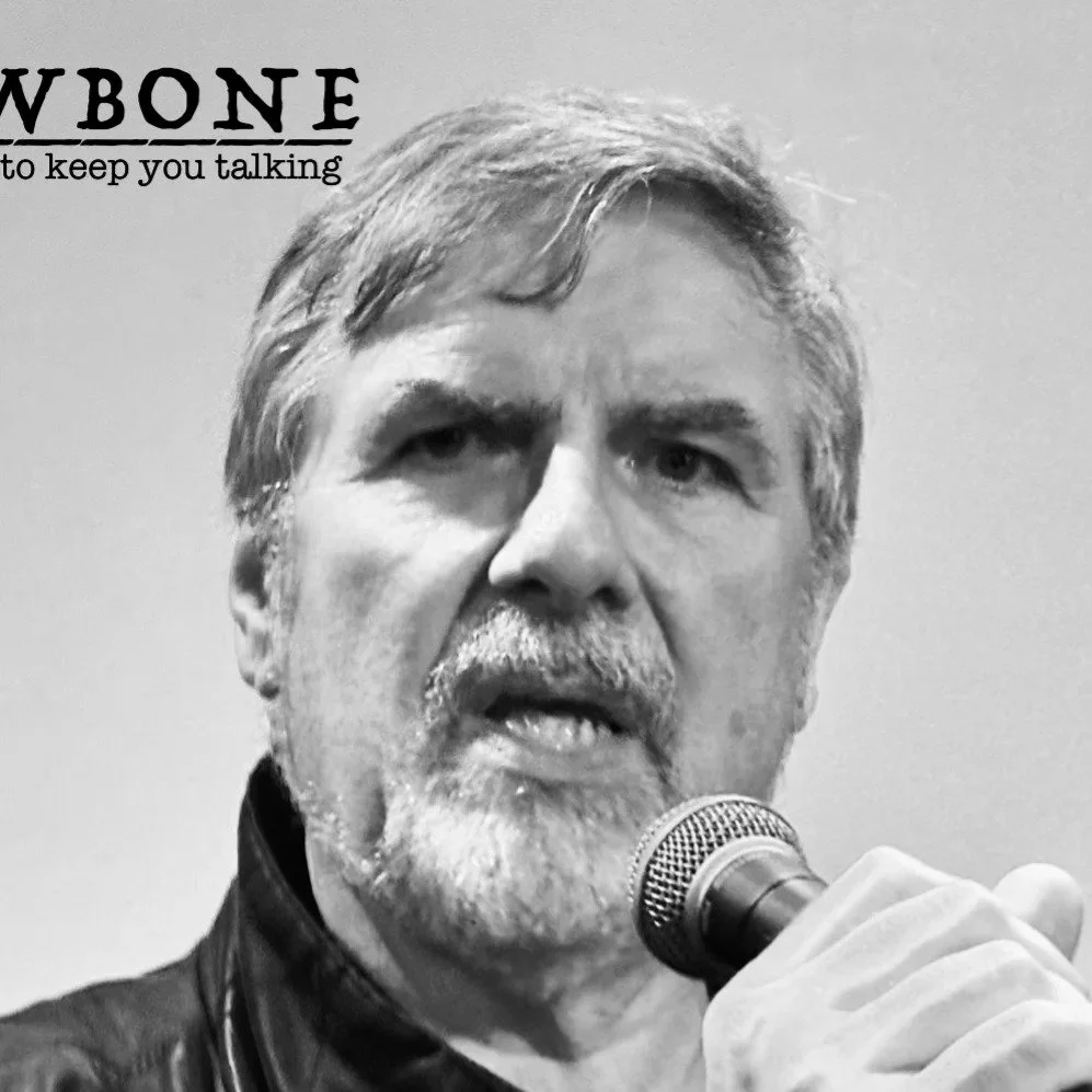 A black and white photo of a man with gray hair and a beard holding a microphone close to his mouth, appearing to speak or sing.