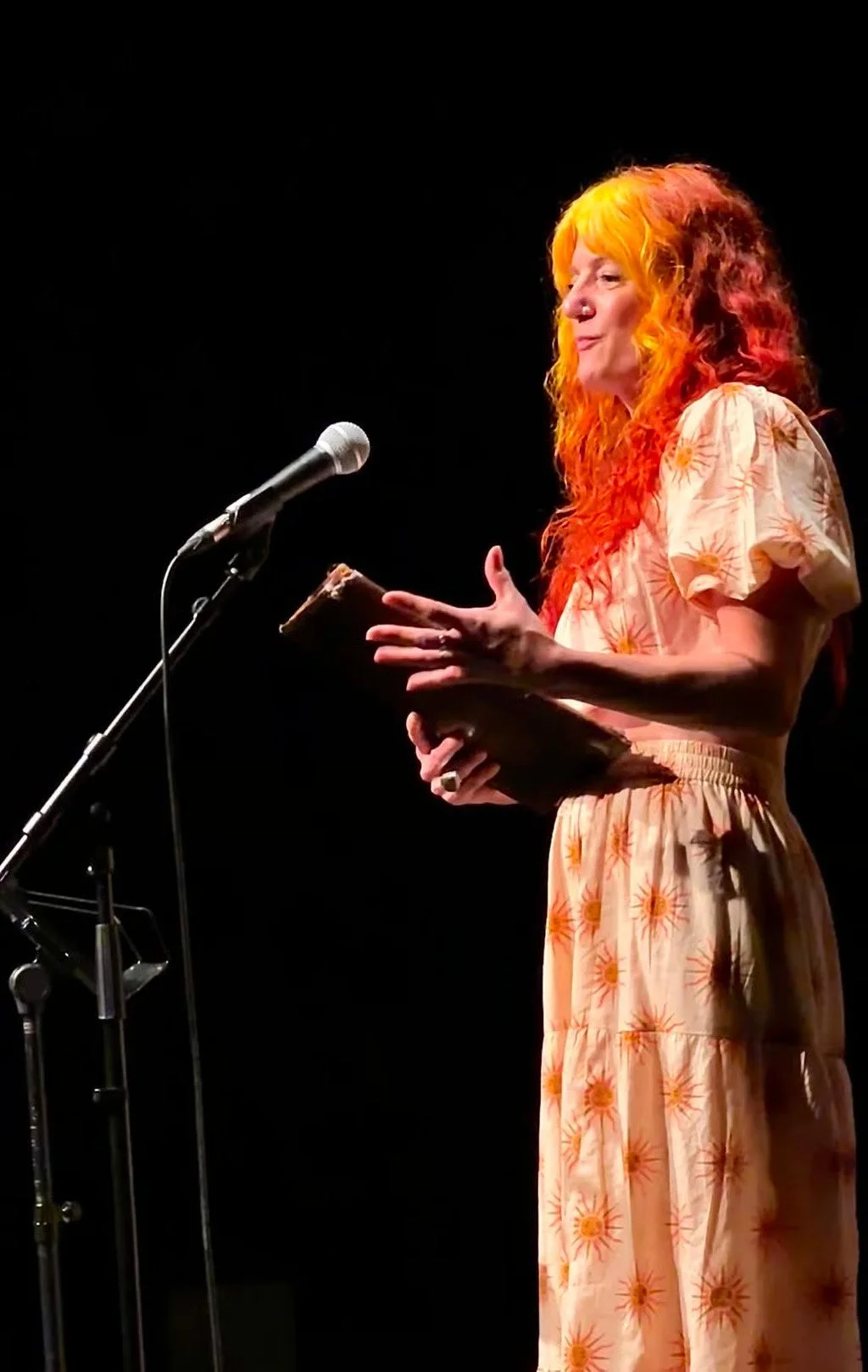 A woman with bright orange hair stands on a dark stage, holding a book, speaking or reading into a microphone.