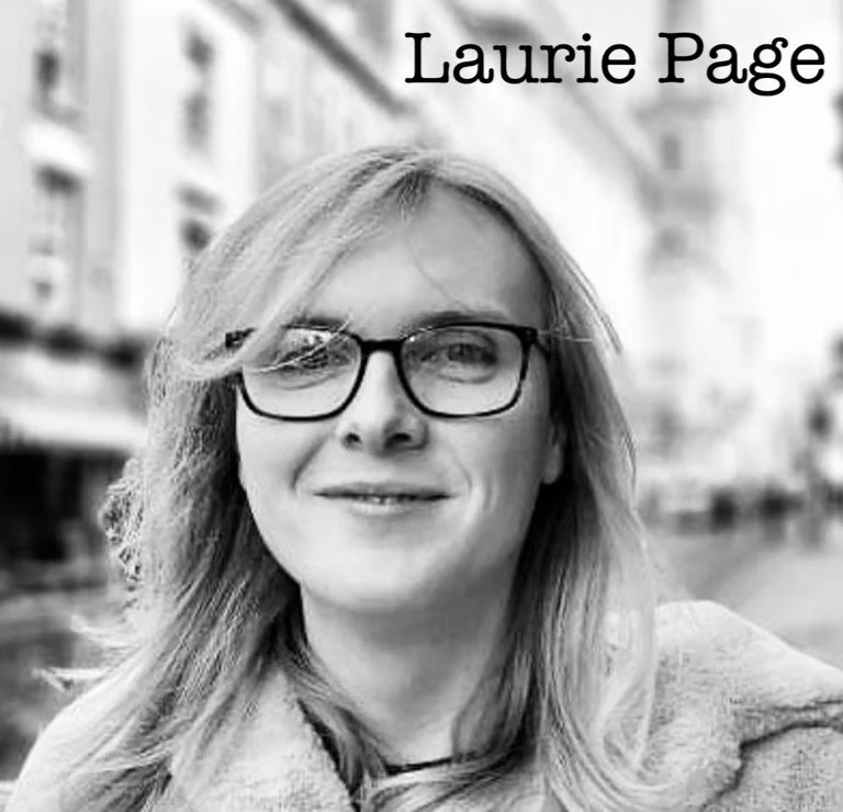 Black and white photo of Laurie Page smiling outdoors in an urban area, wearing glasses and a coat, with her hair blowing in the wind.