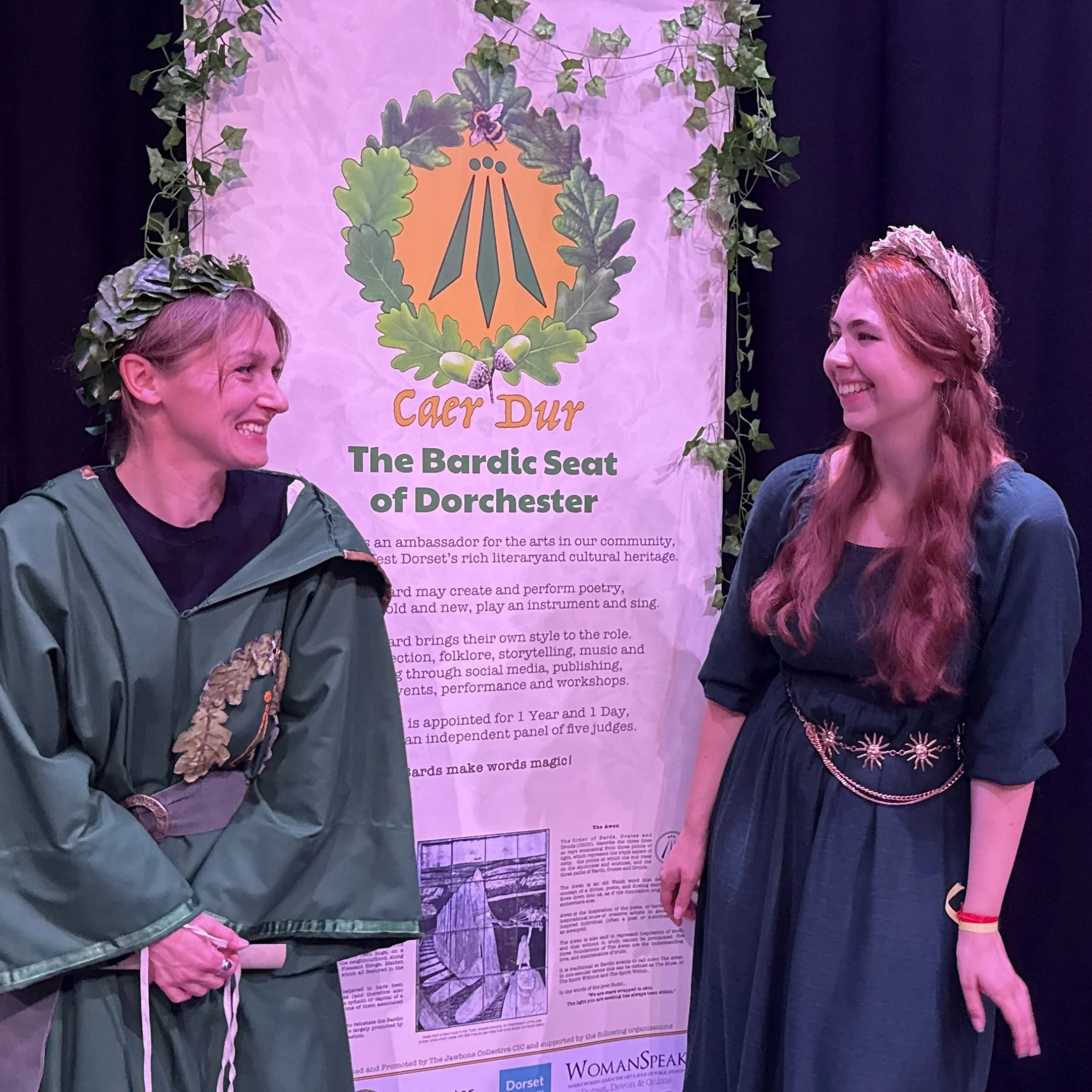 Two women are standing in front of a poster about Caer Ddur, a place in Dorset. One woman has red hair with a floral headband, and the other has short blonde hair and is wearing a green jacket decorated with leaves.