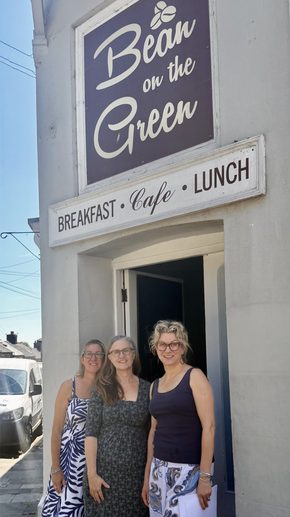Three women standing outside the entrance of a cafe called 'Bean on the Green,' which offers breakfast, cafe, and lunch. The women are smiling and posing for the photo in front of the cafe's door on a sunny day.
