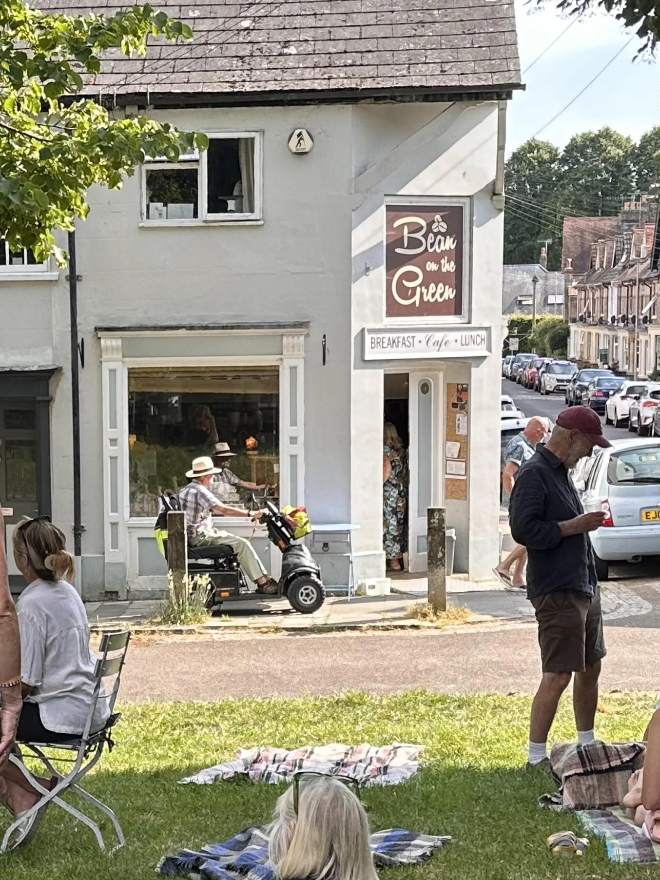 People gathered outside a cafe called 'Bean on the Green' in a small town, sitting and standing on the sidewalk and grass under trees on a sunny day.