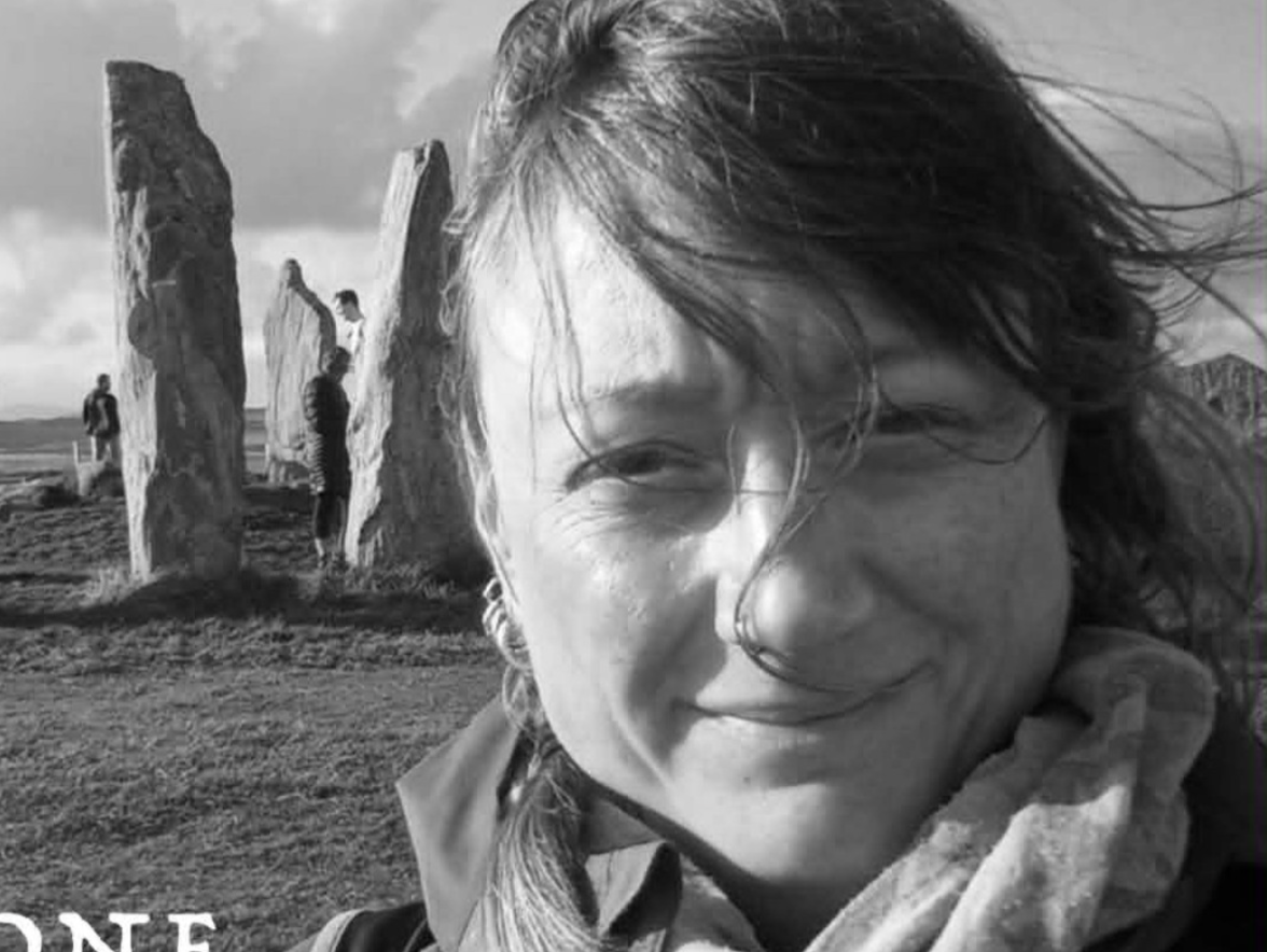 Close-up black and white photo of smiling woman with windblown hair in front of ancient standing stones or monoliths outdoors, with other people visible in the background.